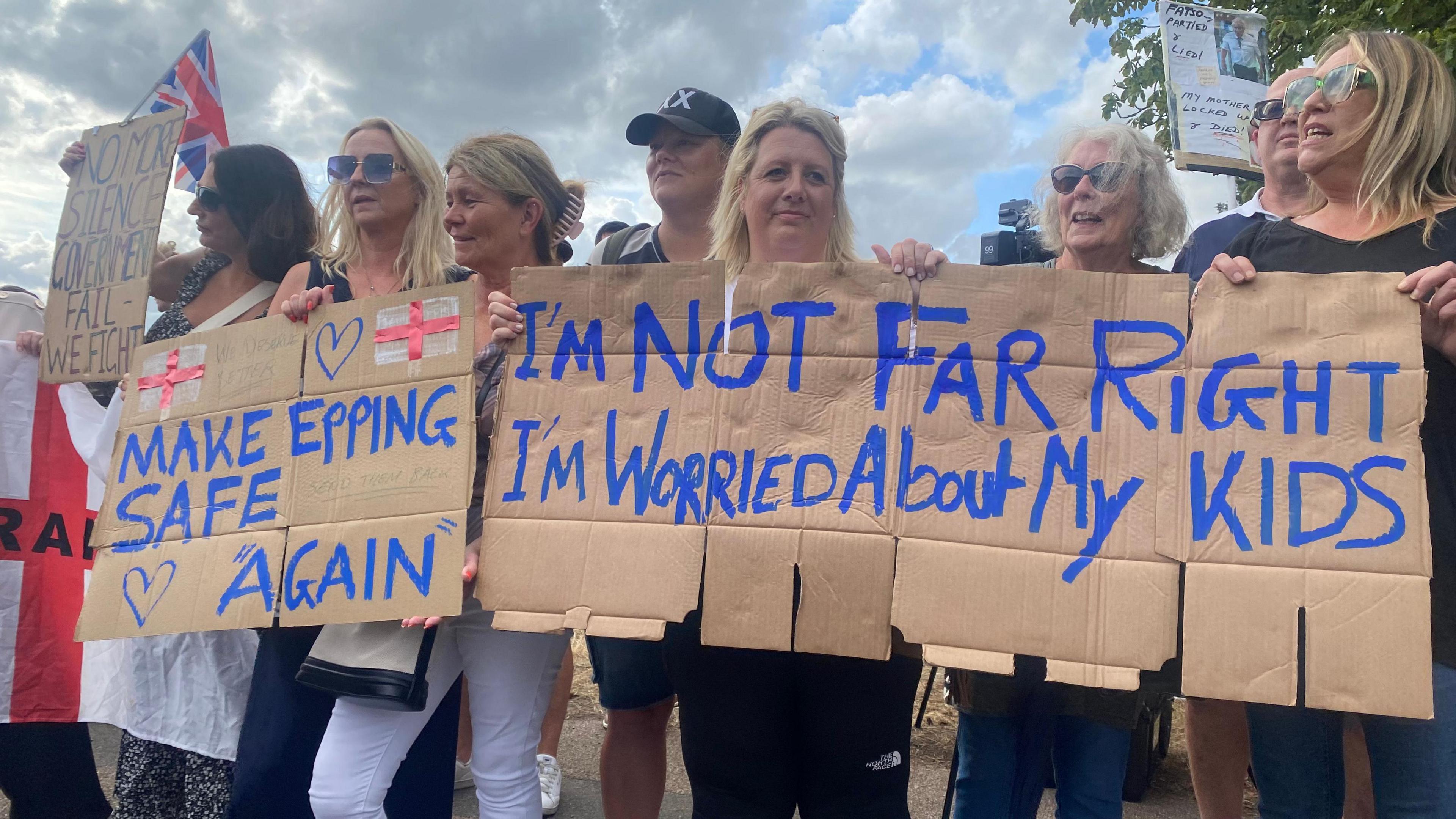 A line of women holding handmade cardboard banners, saying "Make Epping Safe Again" and "I'm not far right, I'm worried about my kids". Some are wearing sunglasses. The banners also bear hearts and England flags