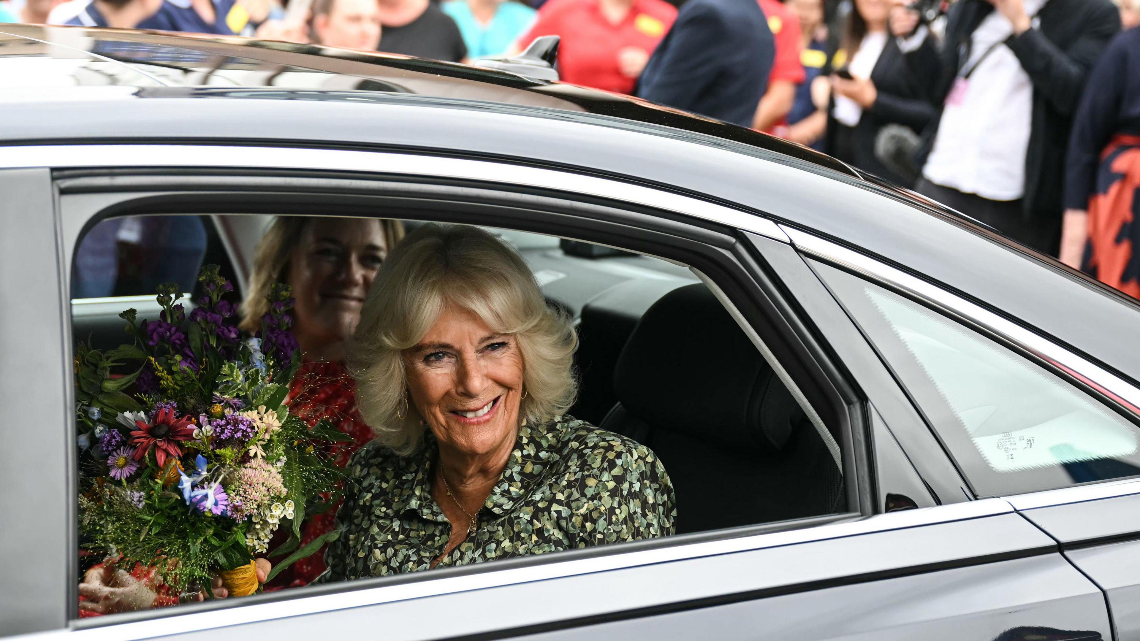 Queen Camilla. She is sat in a black car with another passenger, holding a bouquet of flowers. She is smiling. Crowds of people can be seen in the distance