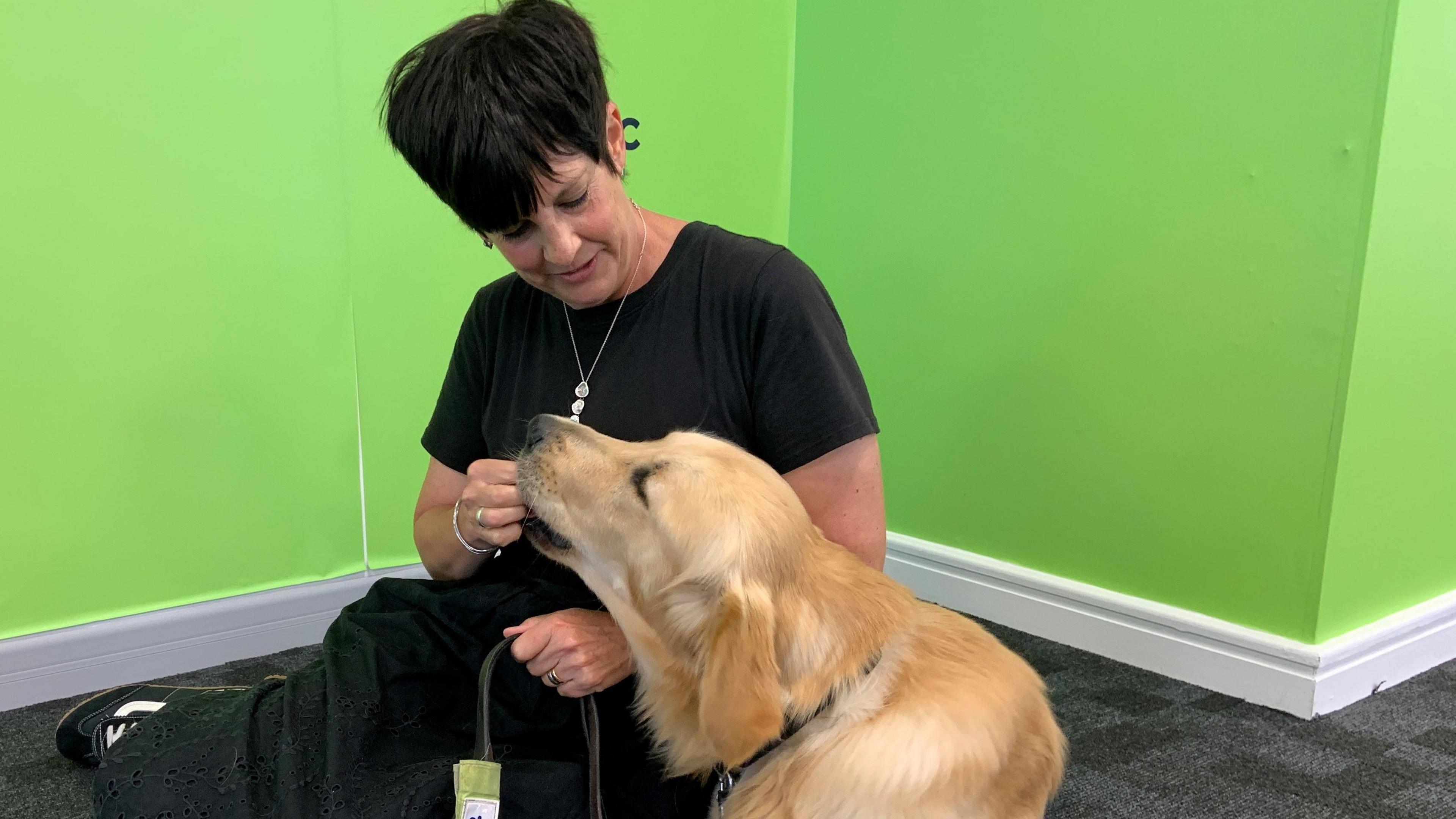 A woman with short black hair is wearing a long black dress. She is sitting on the floor with a golden labrador guide dog and feeding him a treat. There is a bright green wall in the background