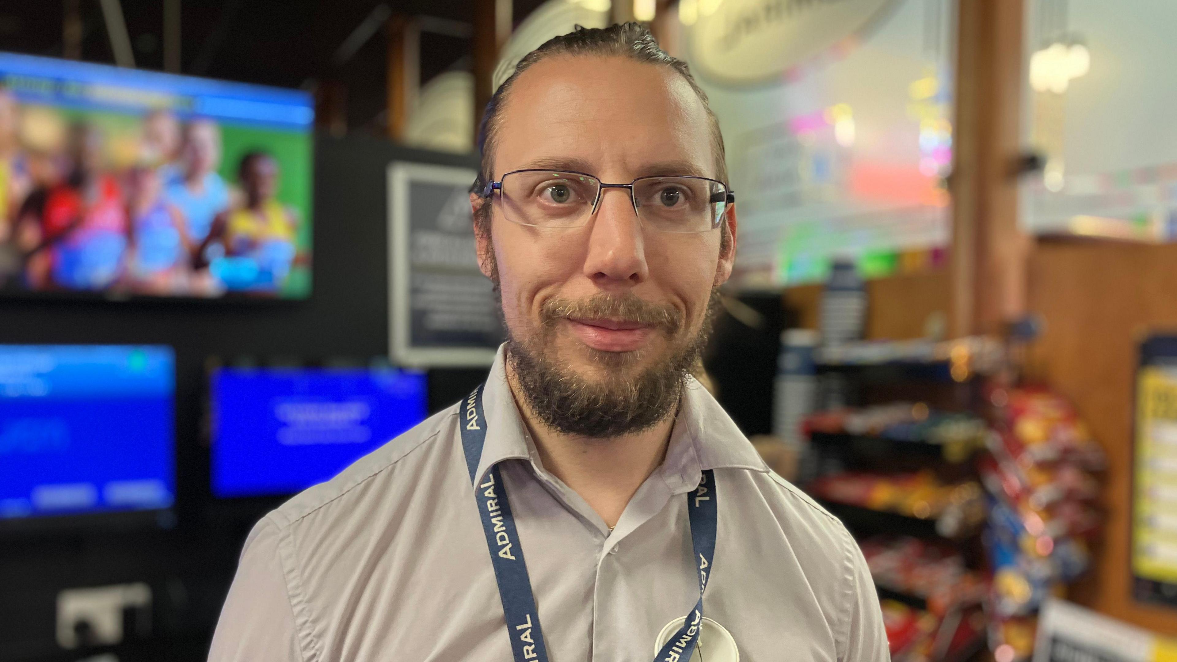 Shane Griffiths - A man with dark brown hair pulled back in a ponytail with a bushy beard smiling, wearing unrimmed rectangle glasses, wearing a grey collared shirt with a blur background