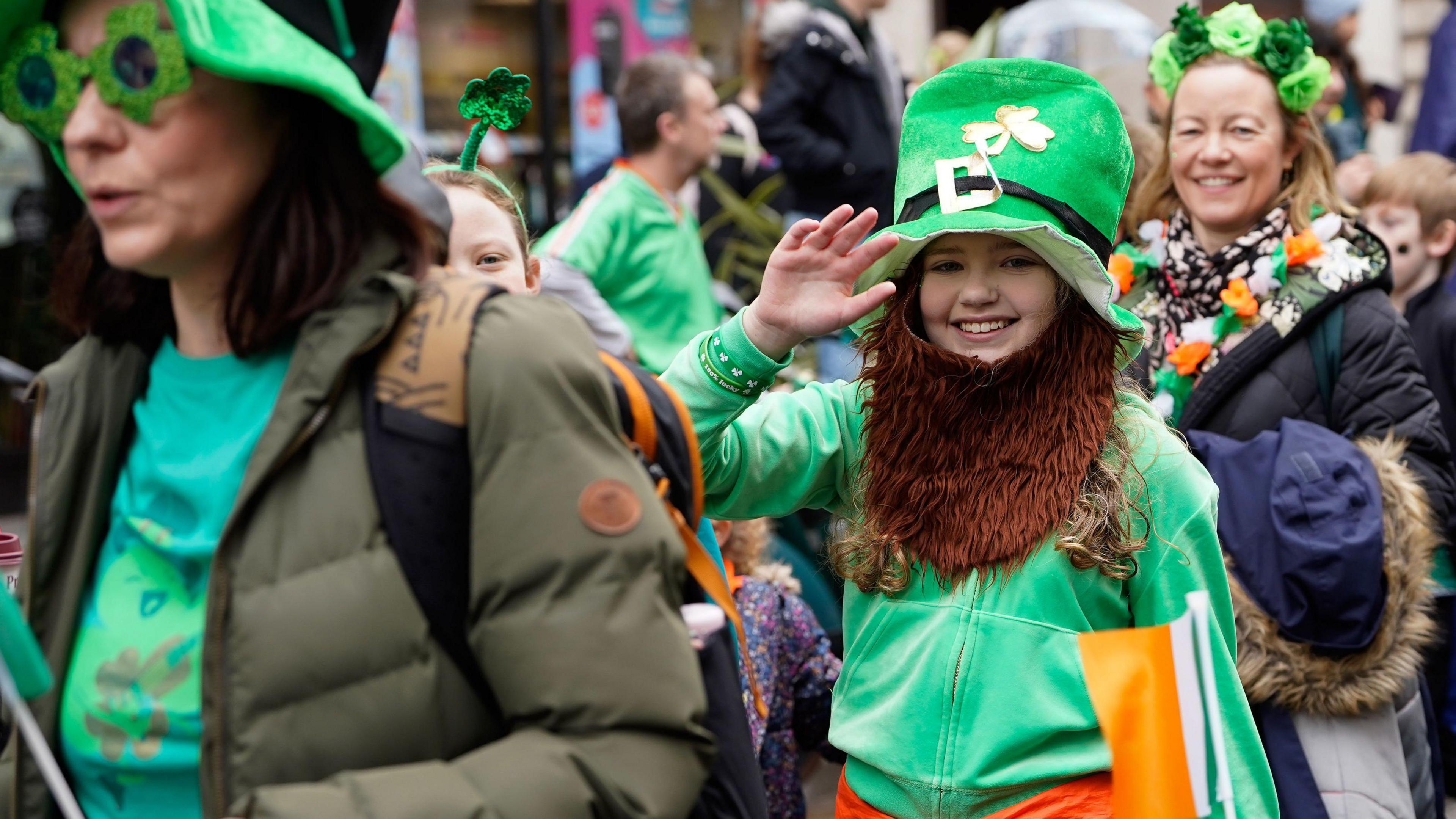 A young girl wearing a green leprechaun costume and ginger beard waves and smiles at the camera while other people wearing green flowers, shamrocks and garlands walk by