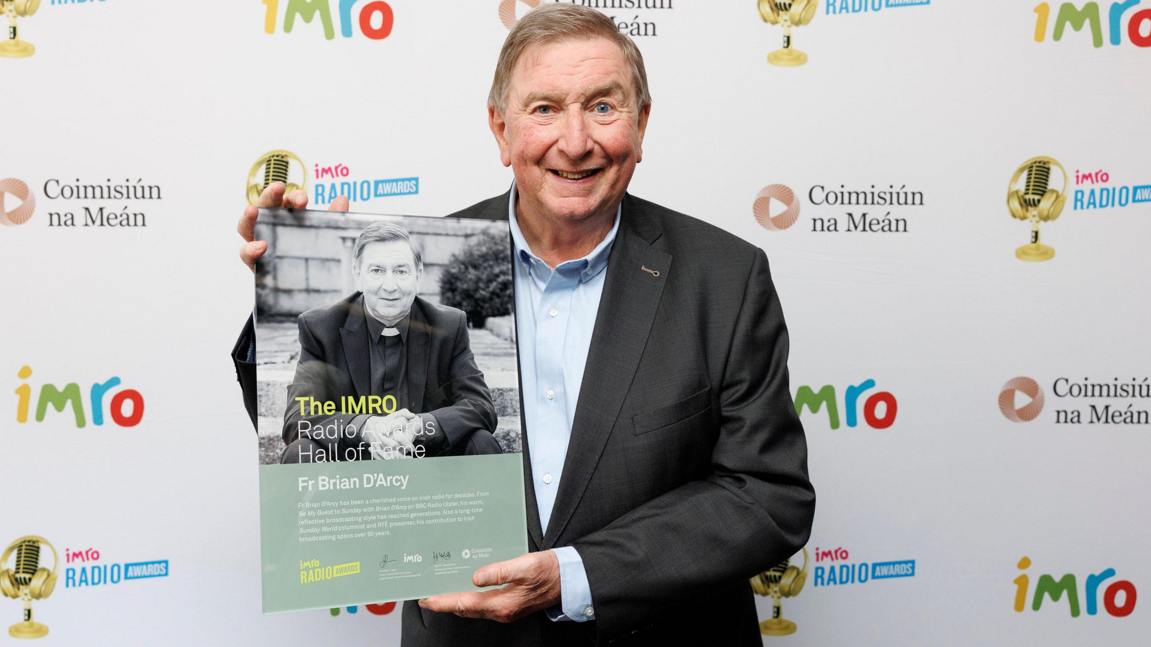 A man with short brown hair smiles at the camera holding a photo of himself. The man wears a black suit and blue shirt. In the photo he wears a black suit, black shirt and a white clerical collar. Behind him is a white board with the words 'IMRO' in yellow, green, blue and red writing. The words 'Coimisiún na Meán' are also on the board in black writing as well as a logo of a gold microphone with the words 'imro Radio Awards.'