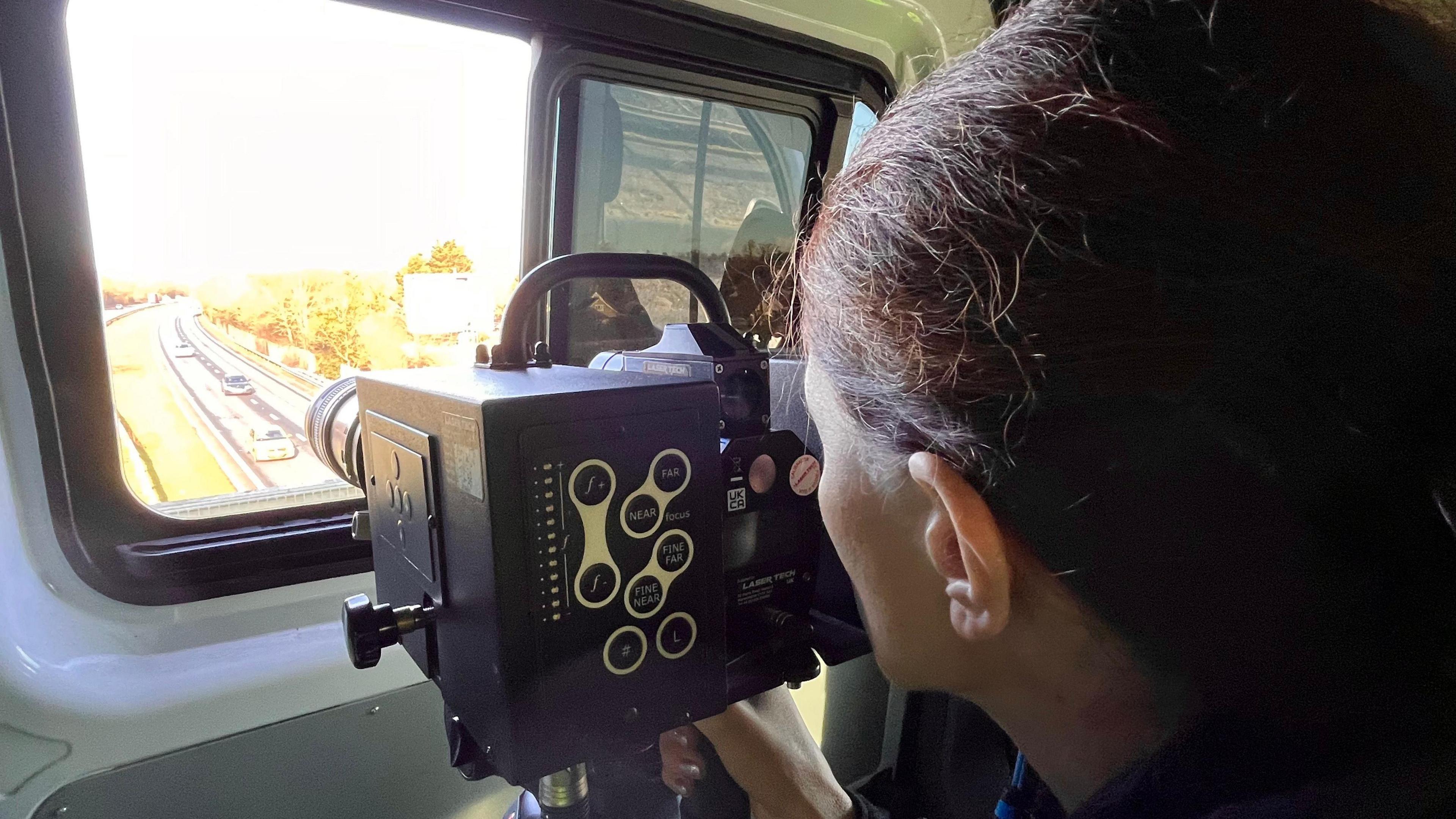 Cara Taylor uses a speed traffic camera to monitor vehicles on the A14 below the bridge her van is parked on. She looks into the camera's viewfinder. Vehicles can be seen travelling on the road below.