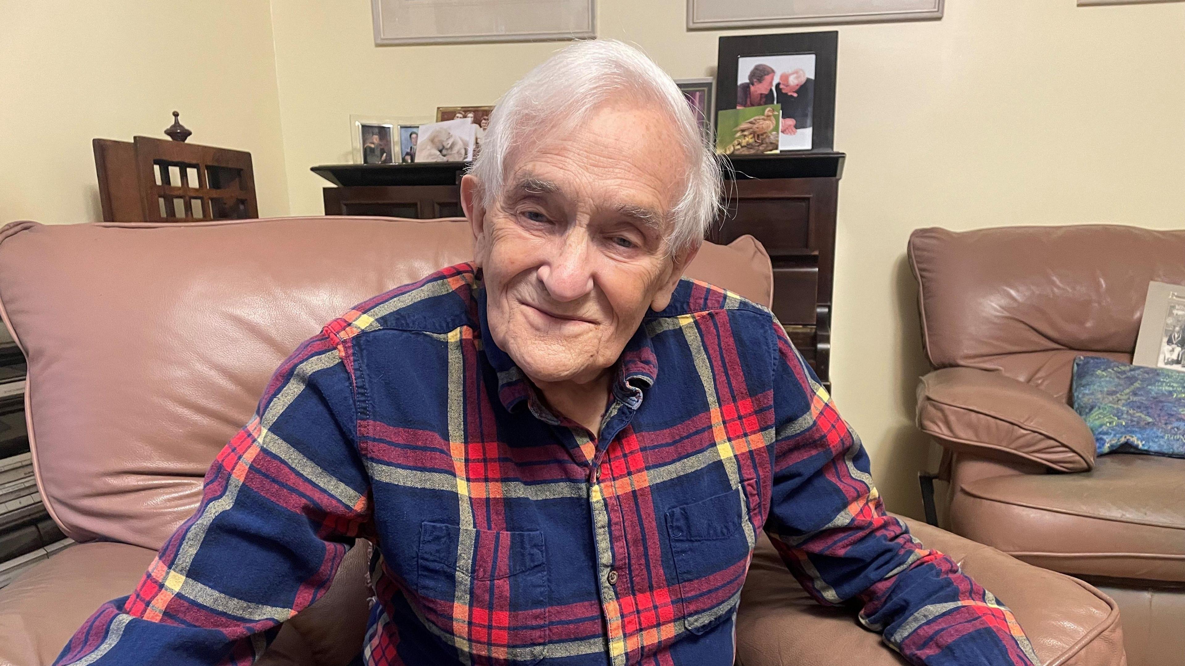 Noel Philo sits in a leather armchair looking towards the camera. He has white hair and is wearing a blue and red panelled shirt