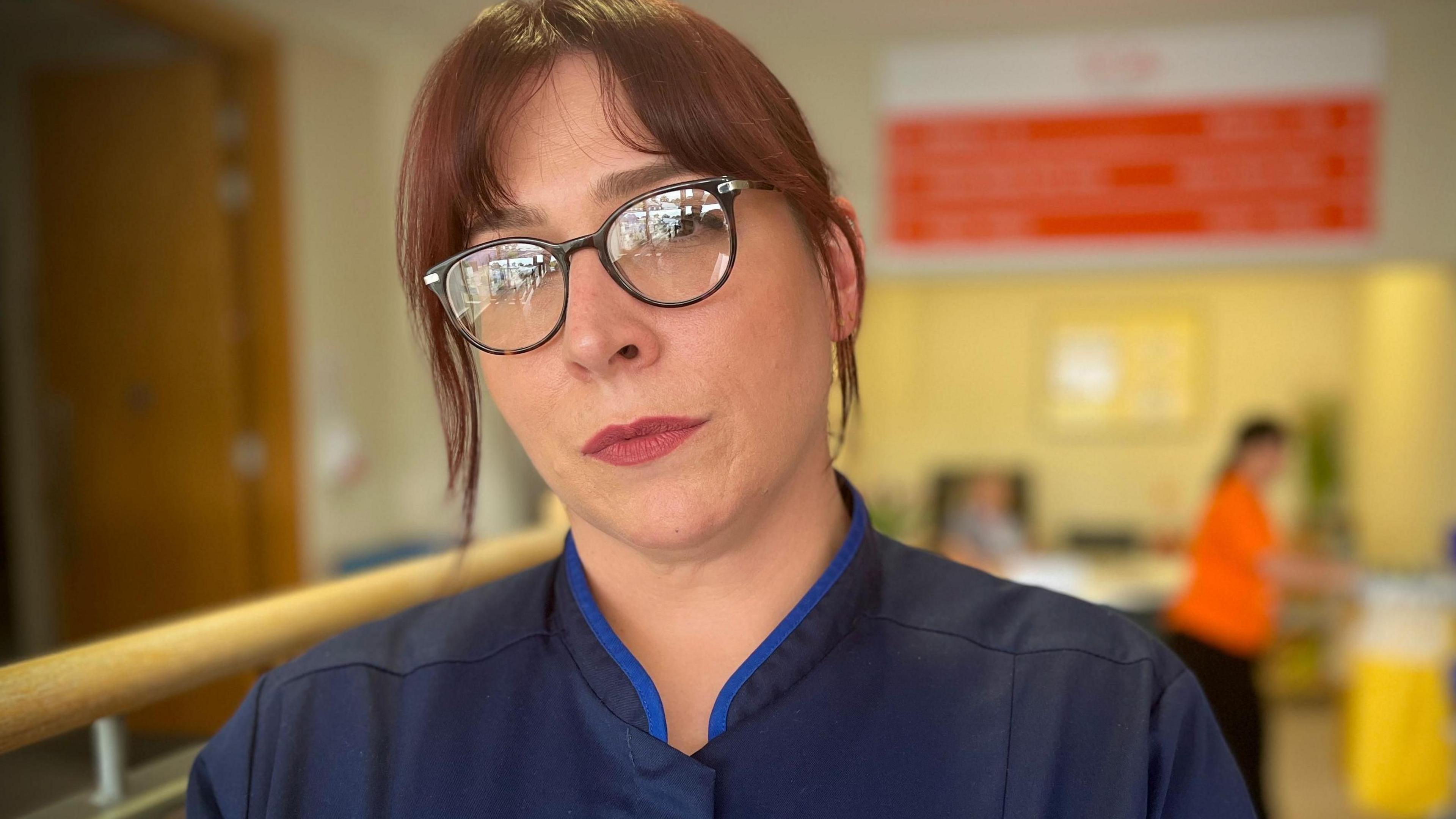 Kristy Clayton looking at the camera. She is wearing glasses and has her hair tied back and is wearing a blue medical top. 