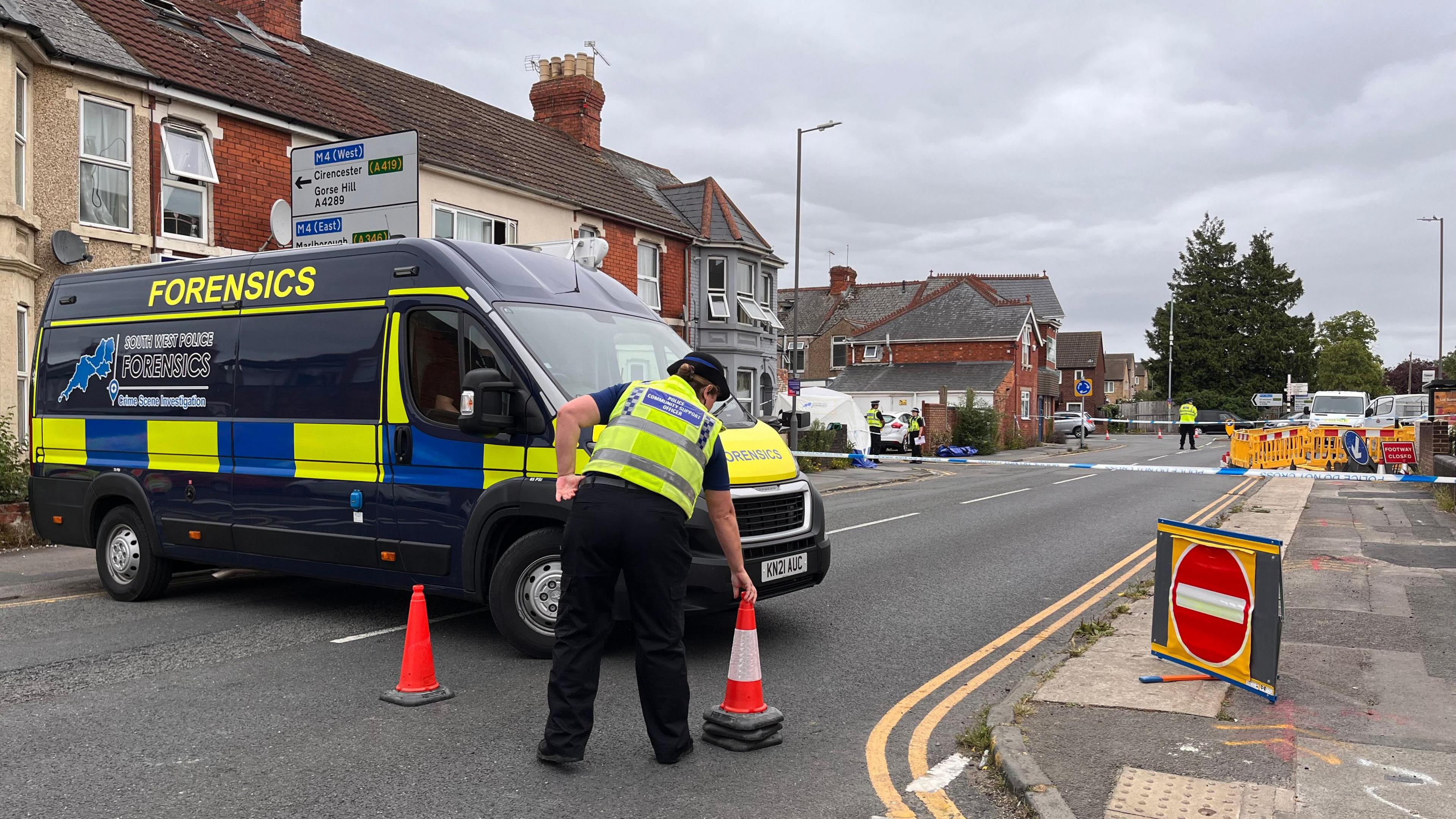 A forensics van with yellow and blue stripes on it. Forensics is written in yellow capital letters at the top. A police cordon can be seen on a residential street, consisting of a red no entry sign and white and blue police tape. An officer is standing next to the van, moving an orange traffic cone. The officer is wearing a yellow high vis jacket with police written on the back.
