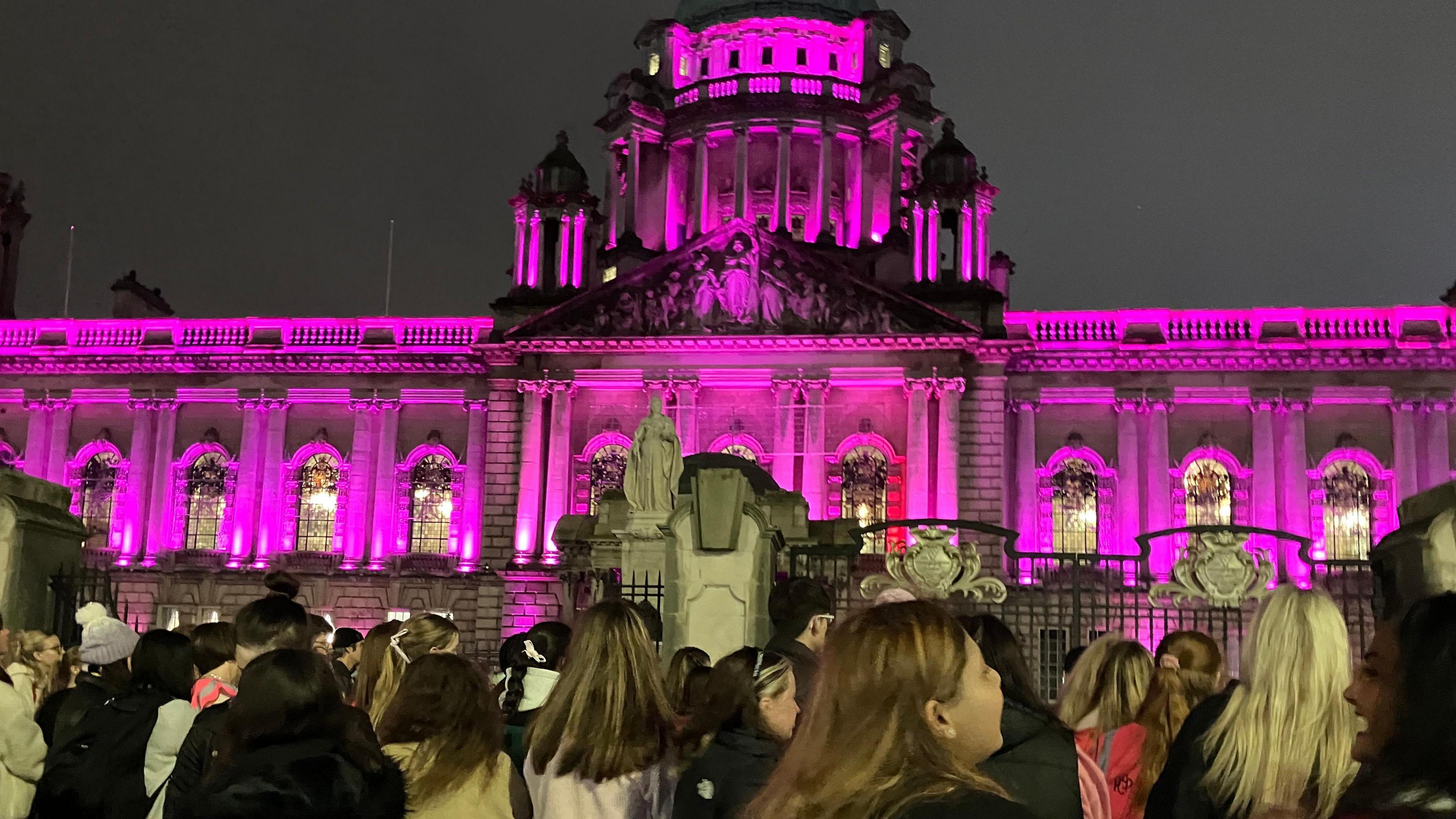 Belfast City Hall is lit up by pink lights. Its night time and 100's of marchers are pictured in front of the building behind its black iron gates