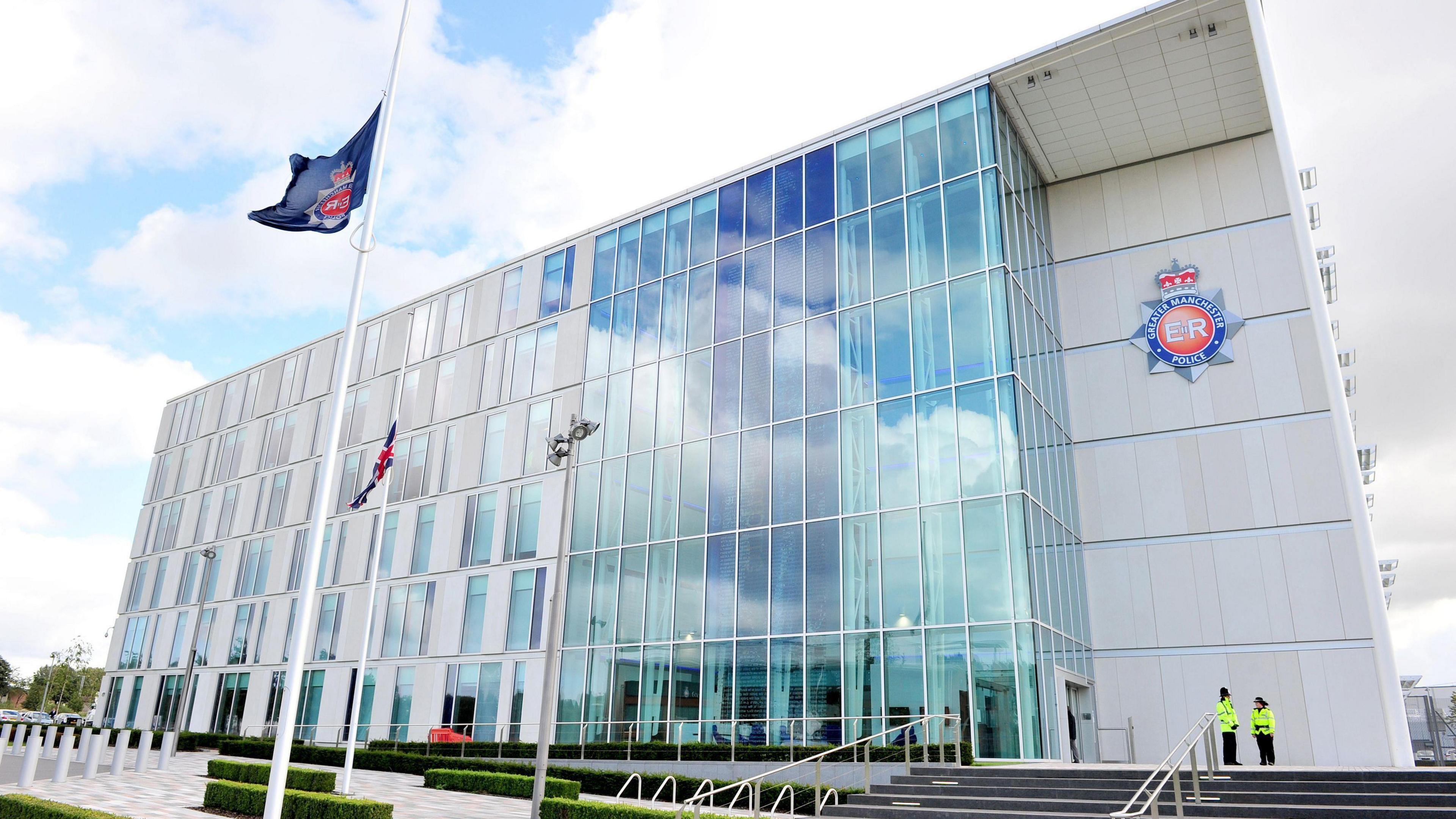 Greater Manchester Police headquarters on a clear day. The building is a multiple-storey building with a glass-panelled front. The force's logo can be seen to one side, with two officers stood on some steps by the entrance. Flagpoles bearing two flag can also been seen in front of the site.