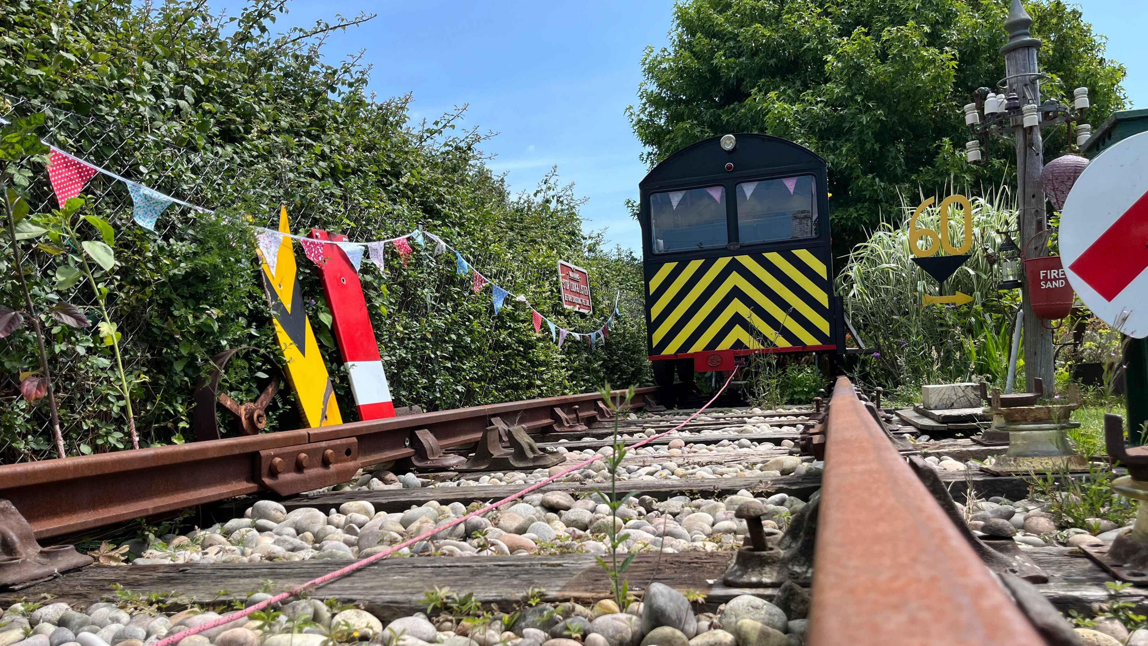 Adrian's train carriage which has yellow and black stripes on the front and is sitting on full sized train tracks. It is pulled by a winch system. Around the edge of the garden are original signs from train lines