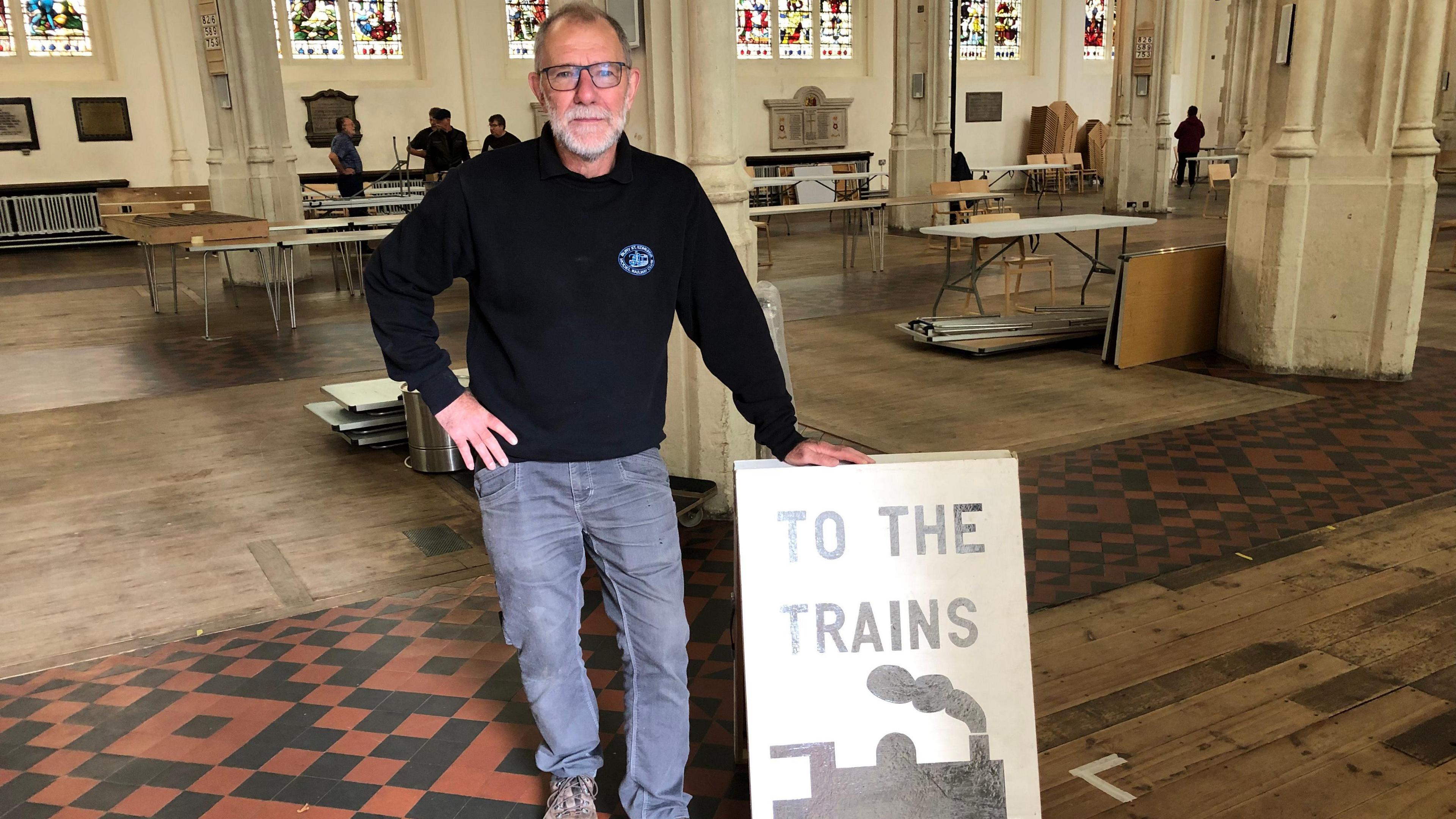 Matthew Porteous is pictured from a wider angle. He is standing next to a sign in the cathedral that reads, 'to the trains'. He has one hand on the sign and another on his hip. Behind him is a part of the cathedral as well as other people.