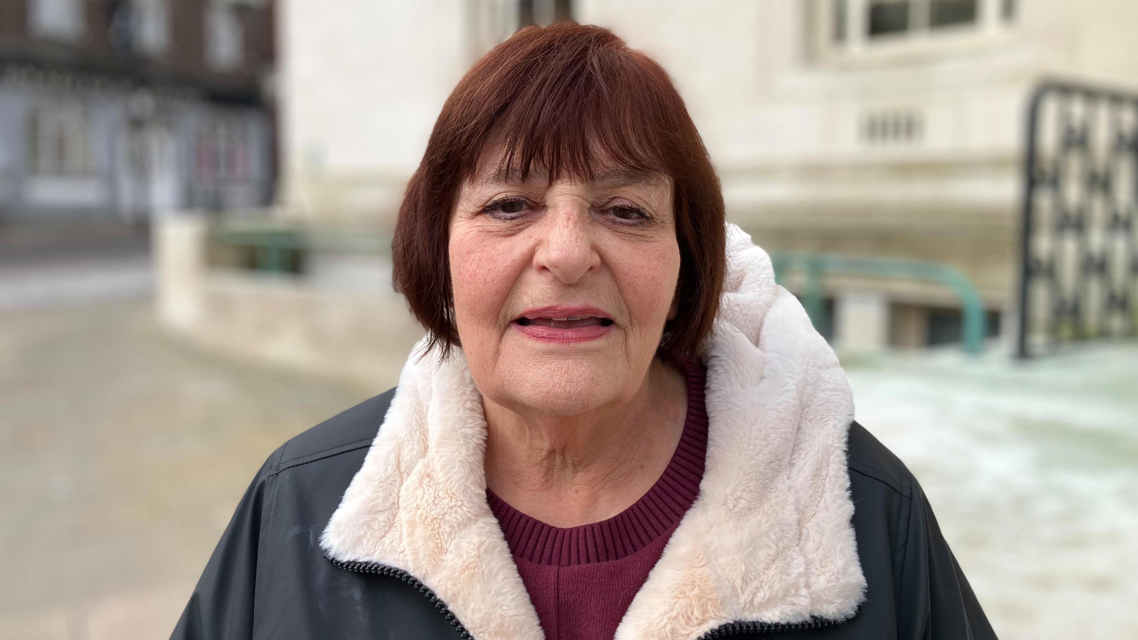 Hazel Simmons stood outside Luton town hall. She has brown hair and is wearing a fur-lined waterproof coat and dark red jumper. She is looking directly at the camera. 