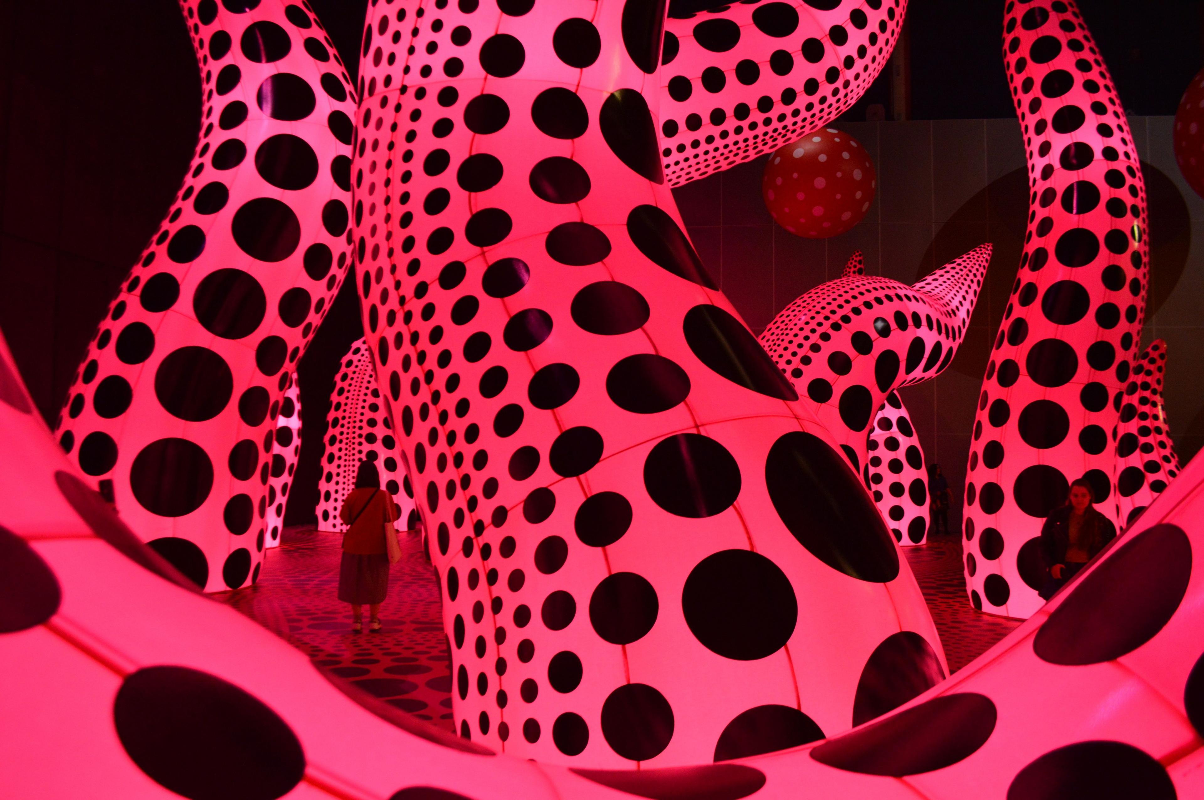 Giant luminous pink tentacle-like sculptures covered in black spots in a dark gallery, with visitors dwarfed by their scale