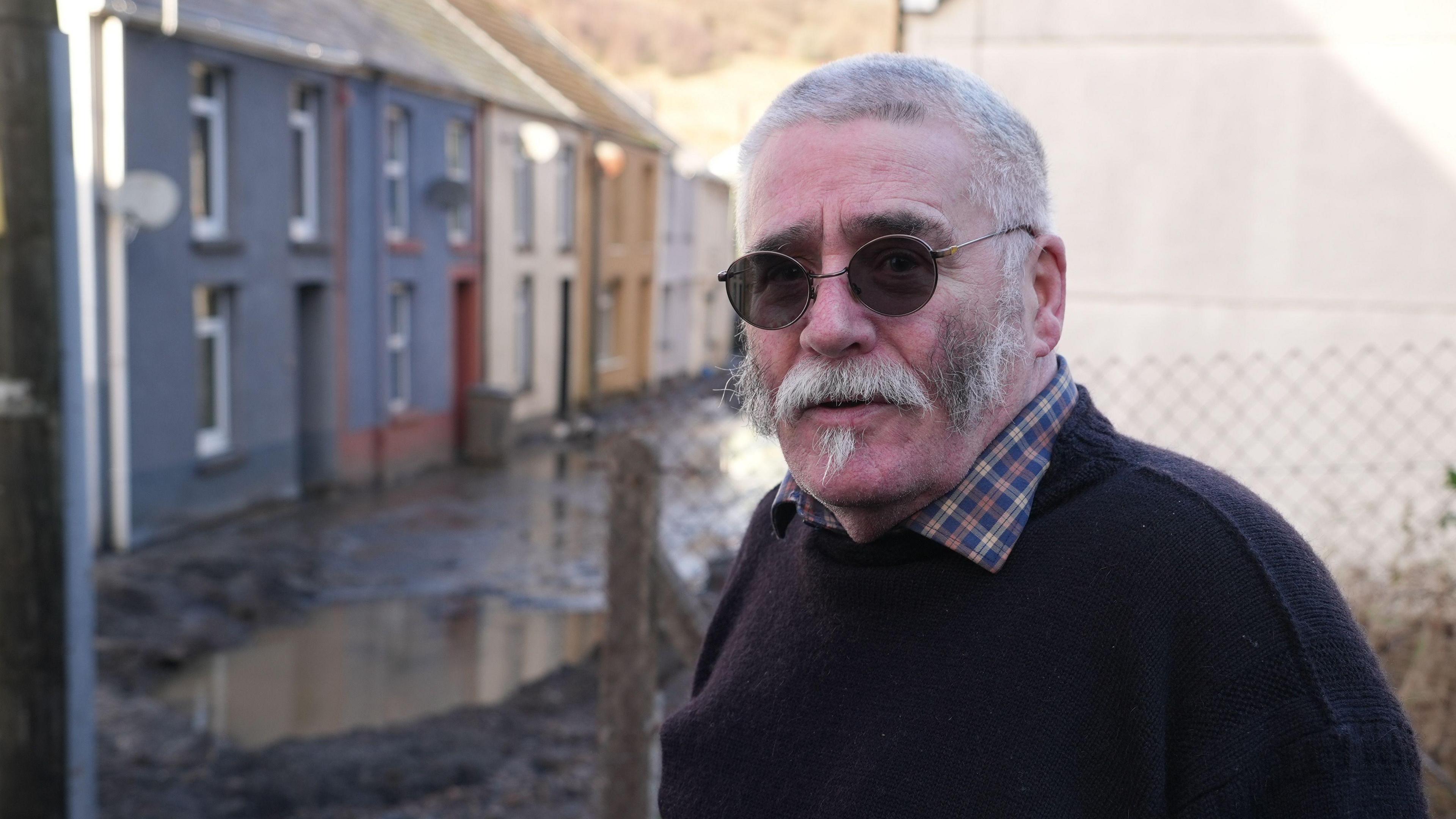 Rob Scholes pictured to the side of a street of terraced houses in front of a wired fence. The road is covered in mud and water. He is wearing a navy jumper and checked shirt and glasses, and has a moustache and short grey hair.