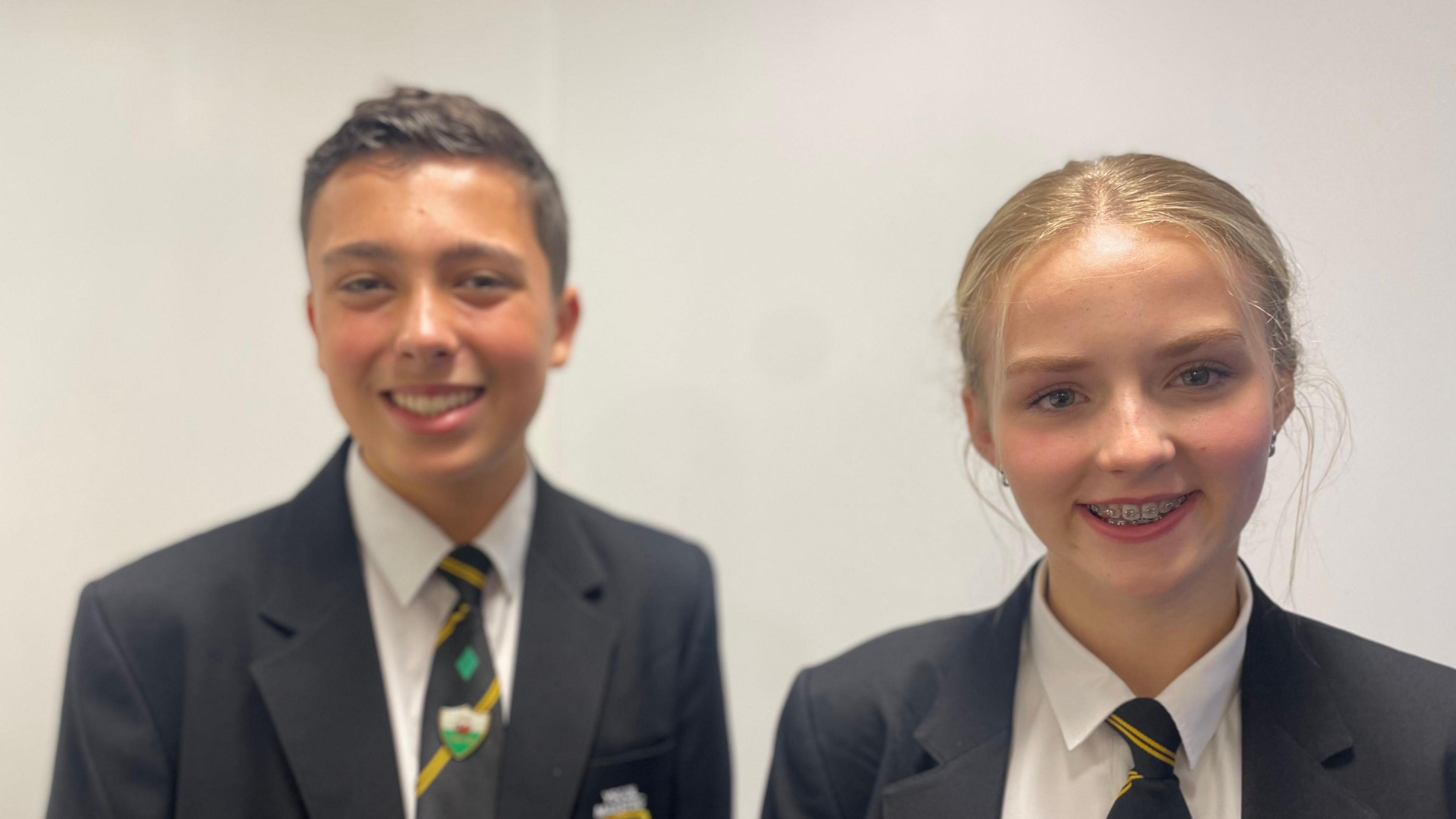 Two pupils in school uniform - which is a black blazer, a black tie with diagonal yellow stripes and a white shirt - standing in front of a plain white wall. The boy on the left, with short dark hair, has a Welsh flag badge on his tie and the girl on the right has blonde hair tied back.