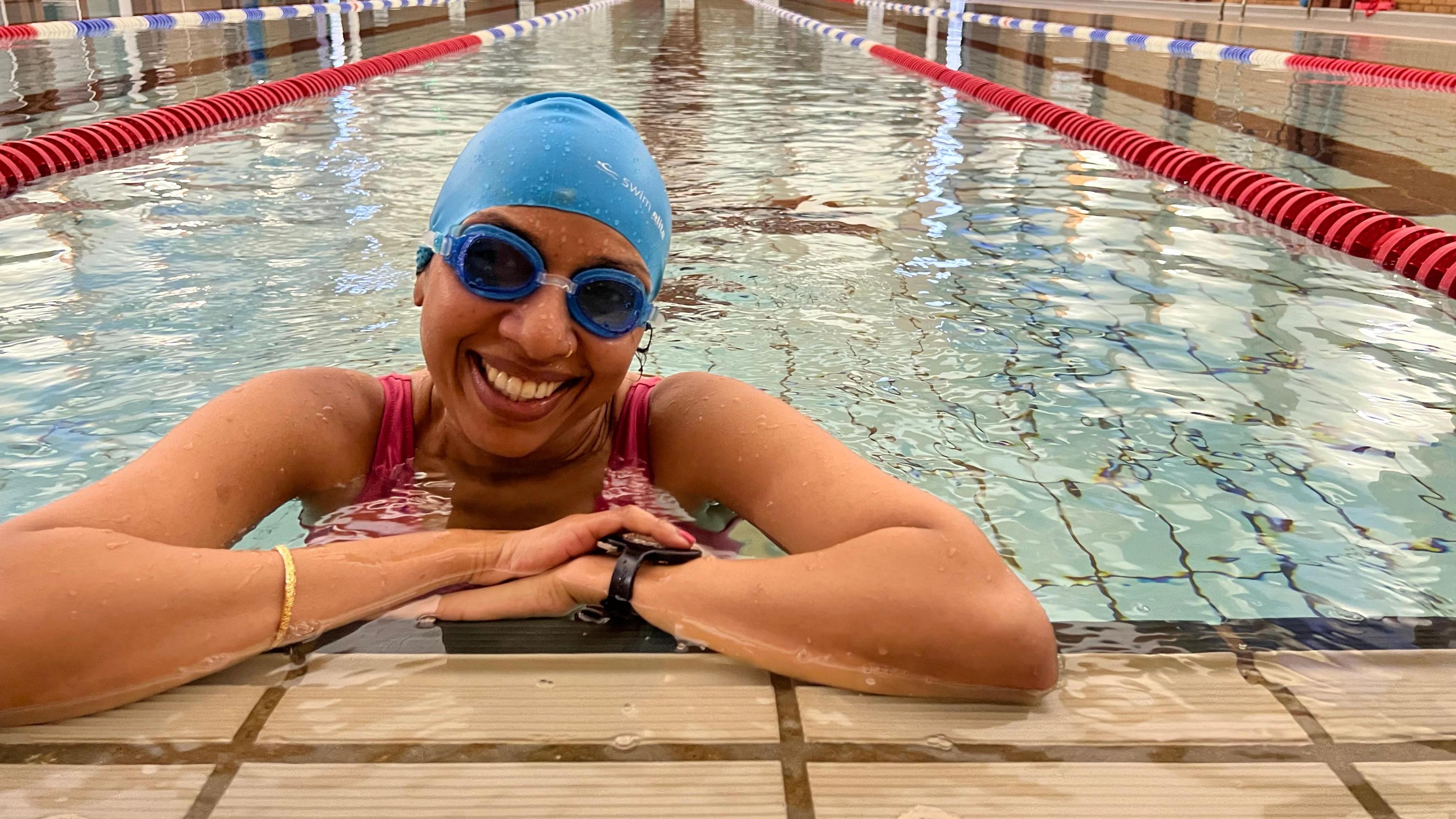 A woman in a pool, resting her elbows on the edge, smiling at the camera wearing a blue swimming hat and blue goggles.