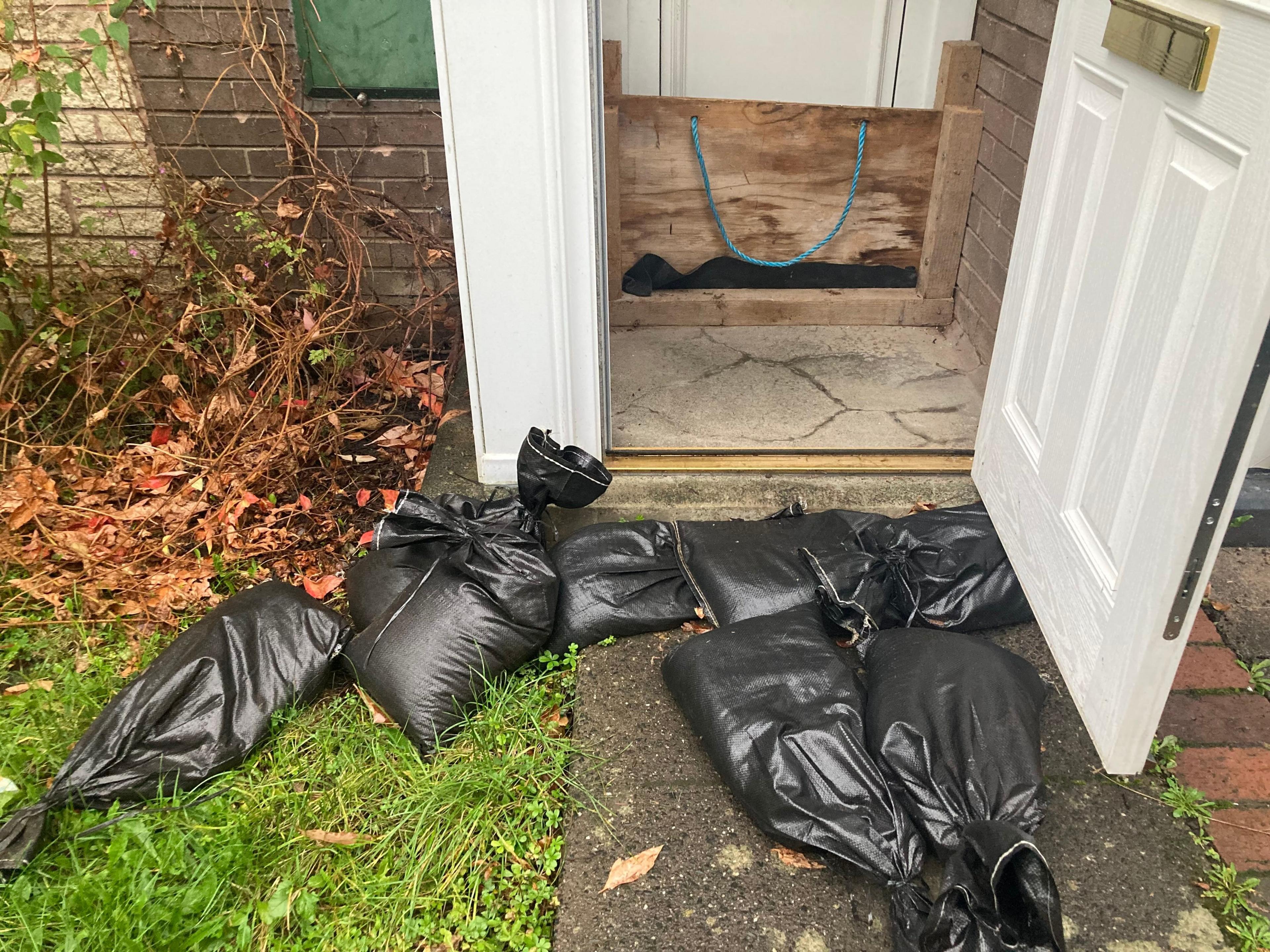 Six black plastic sandbags life on the ground in front of the front door of a house. The door is ajar and on the inside is a small wooden barrier