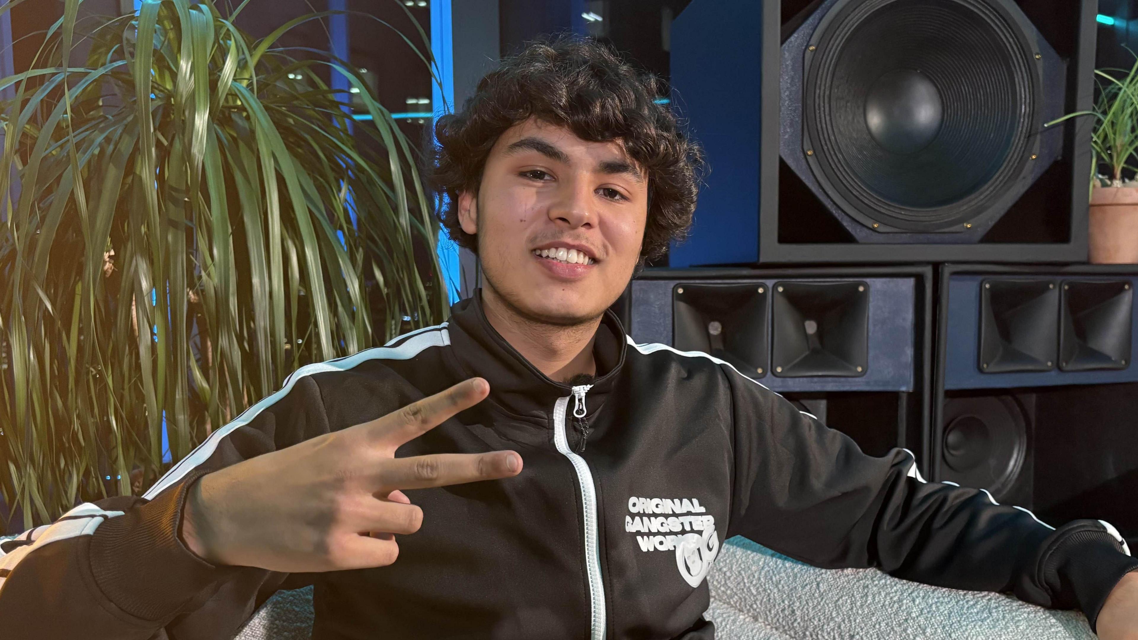 Haydog sits in a white cushioned chair. He holds up a V sign with his right hand. He's wearing a zipped up black shell top and grey trousers. There's a plant and a big speaker in the background. 