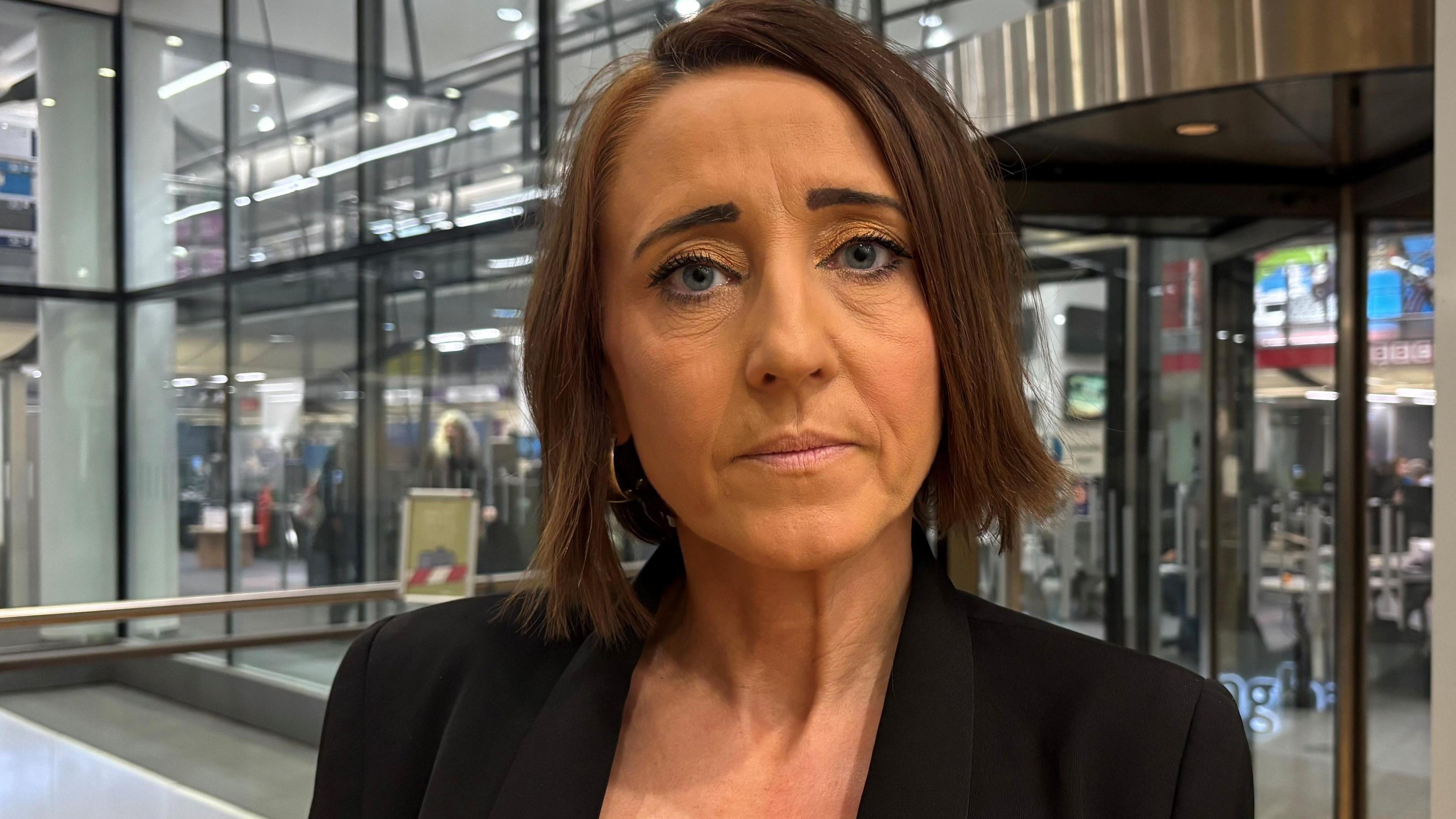 A woman with short dark hair in a black jacket standing in front of glass doors looks directly at the camera 