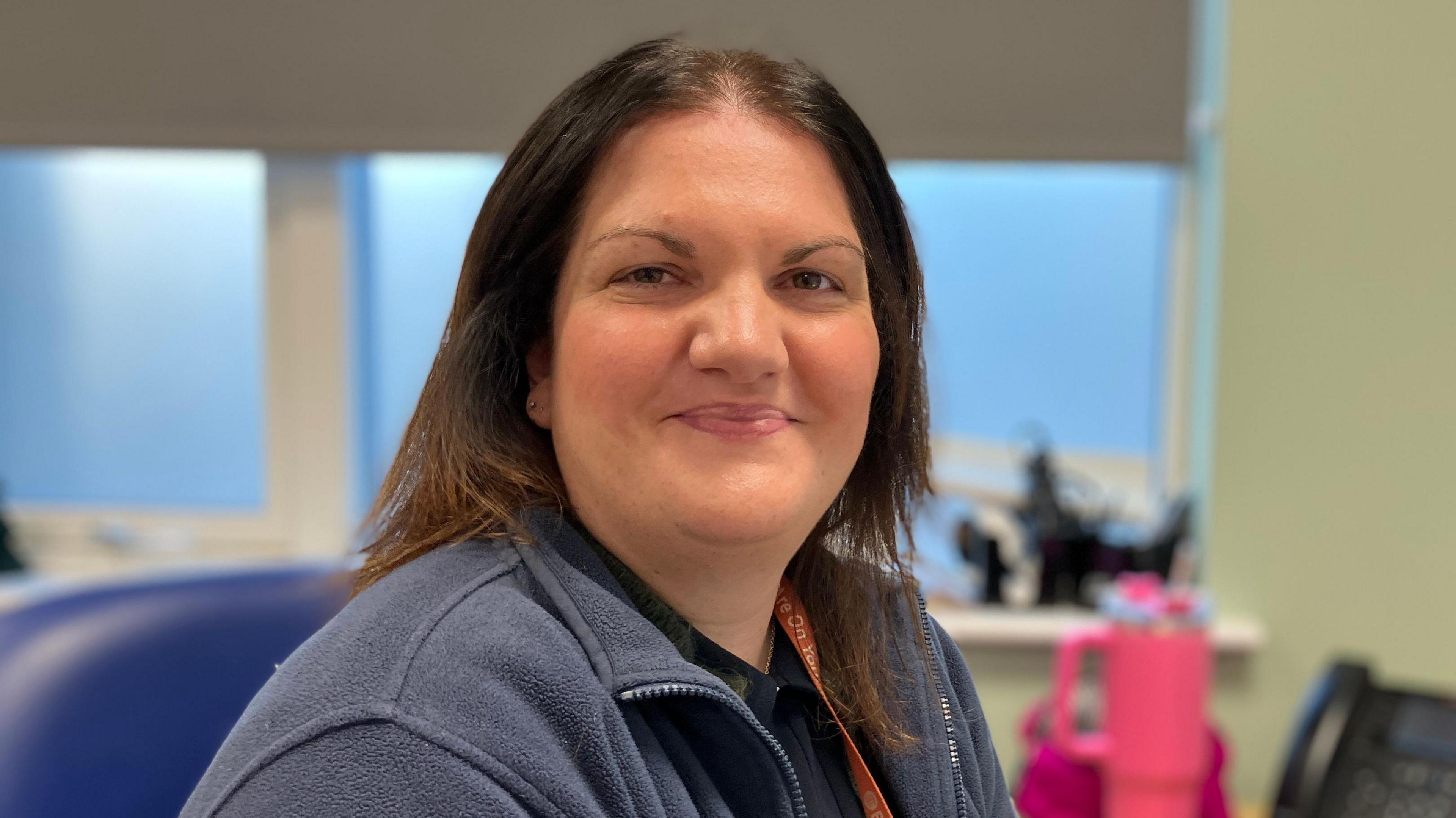  Sarah Sewell, with brown bob-length hair, wearing a blue navy fleece, sits at a desk in an office. 