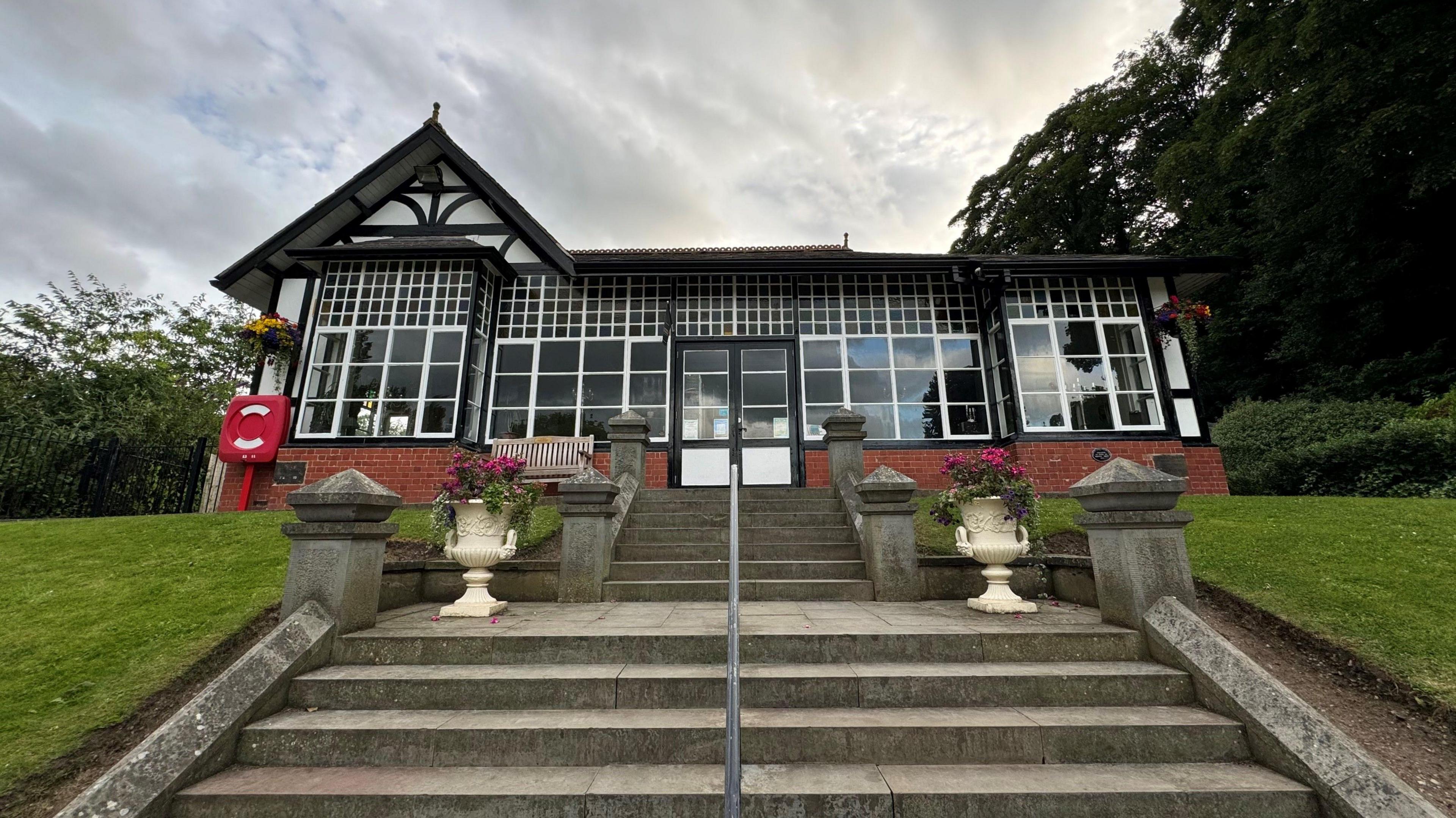 A black and white half-timber building at the top of two flights of steps. It is set within a park, with a lawned embankment in front and trees behind.