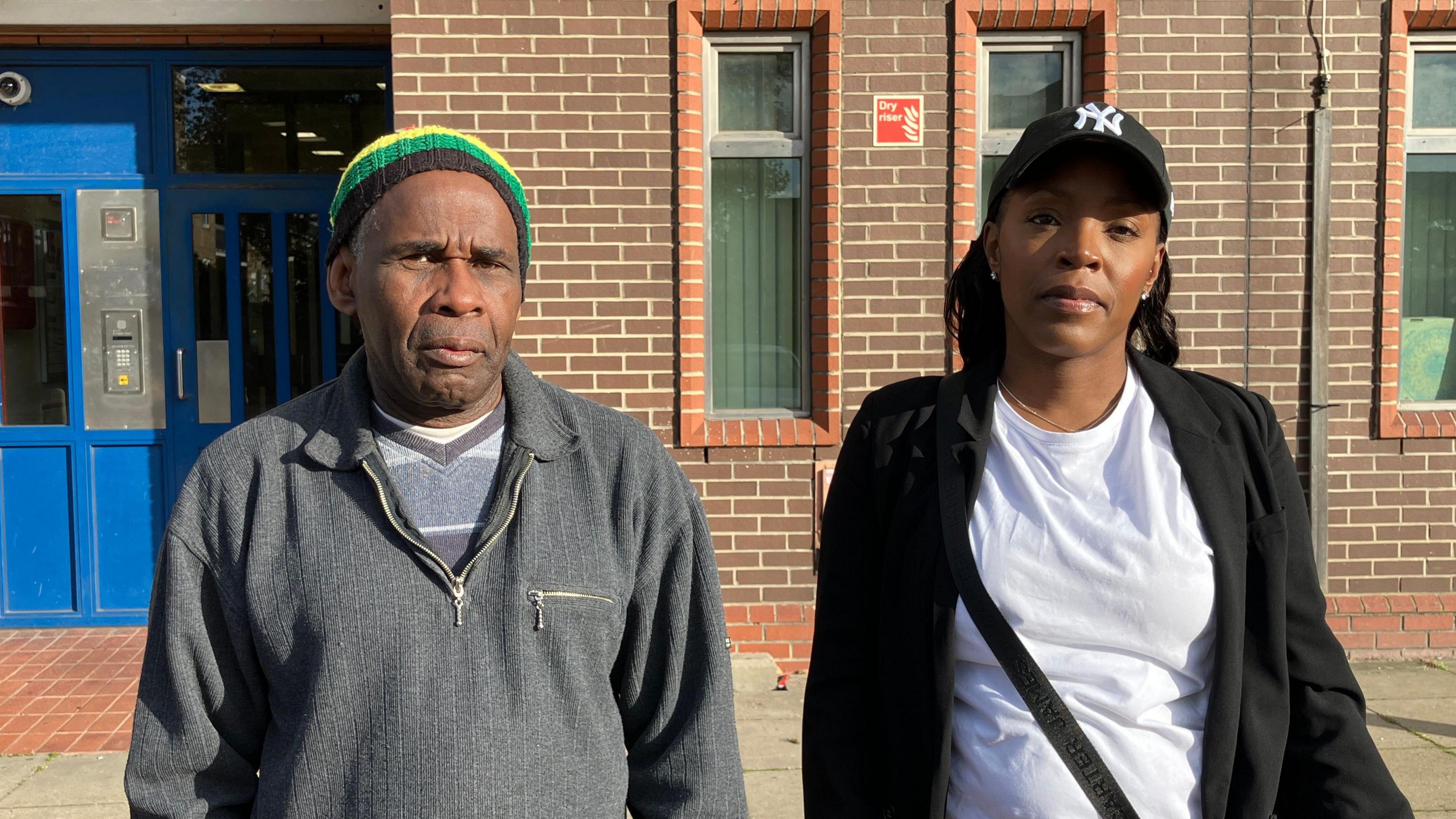 Lesta Hall, a man wearing a red, yellow, blue and black beanie and grey sweatshirt, and Rochelle Wright-Clarke, a woman with long dark hair wearing a black and white baseball cap, white t-shirt and black blazer. They are standing in front of the entrance to Walford House which has blue double doors, a black and white sign and brick walls