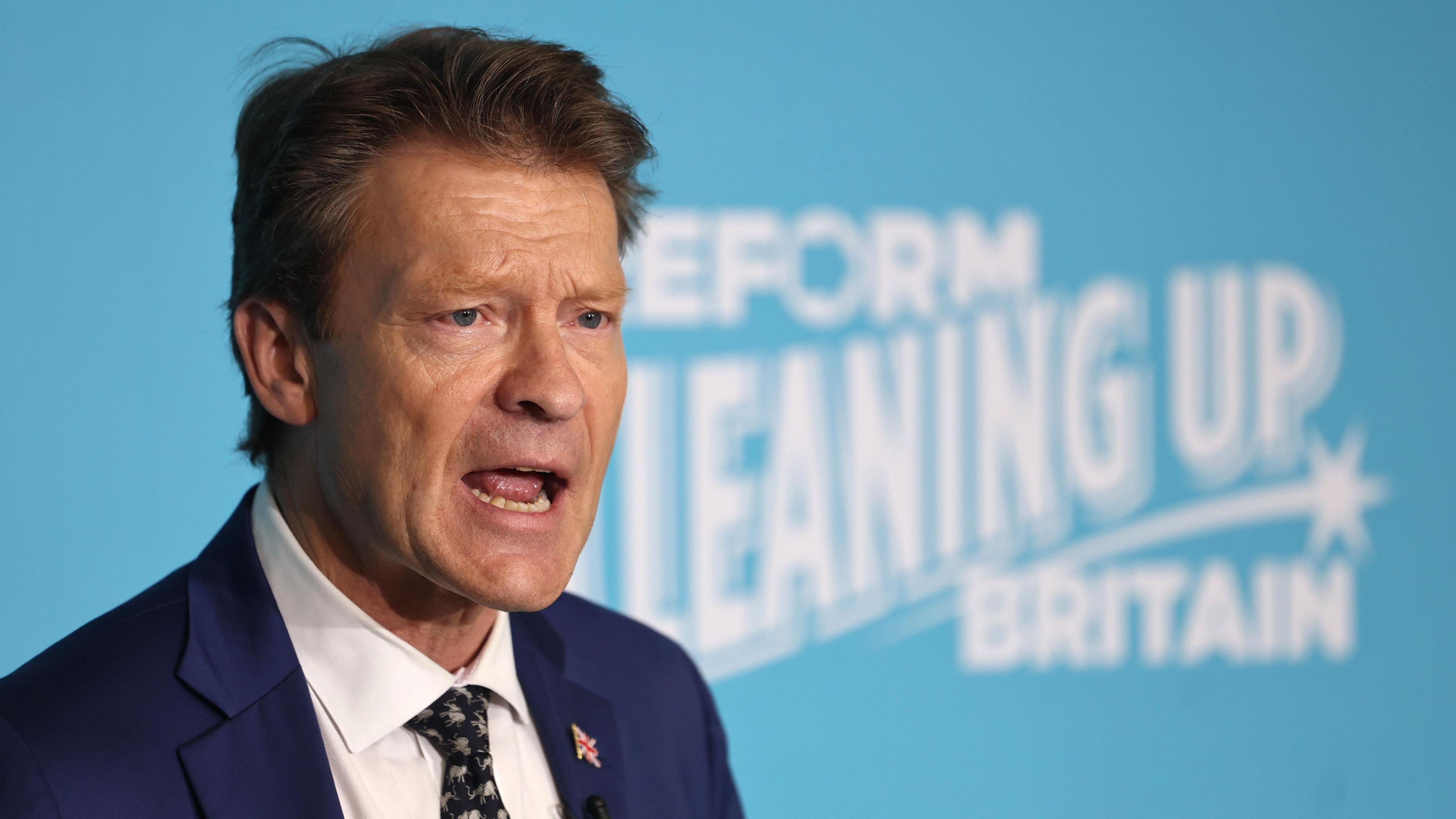 Richard Tice in a dark suit, wearing a union flag button on his lapel. He is standing behind a large turquoise blue sign with white writing which reads: Reform cleaning up Britain 