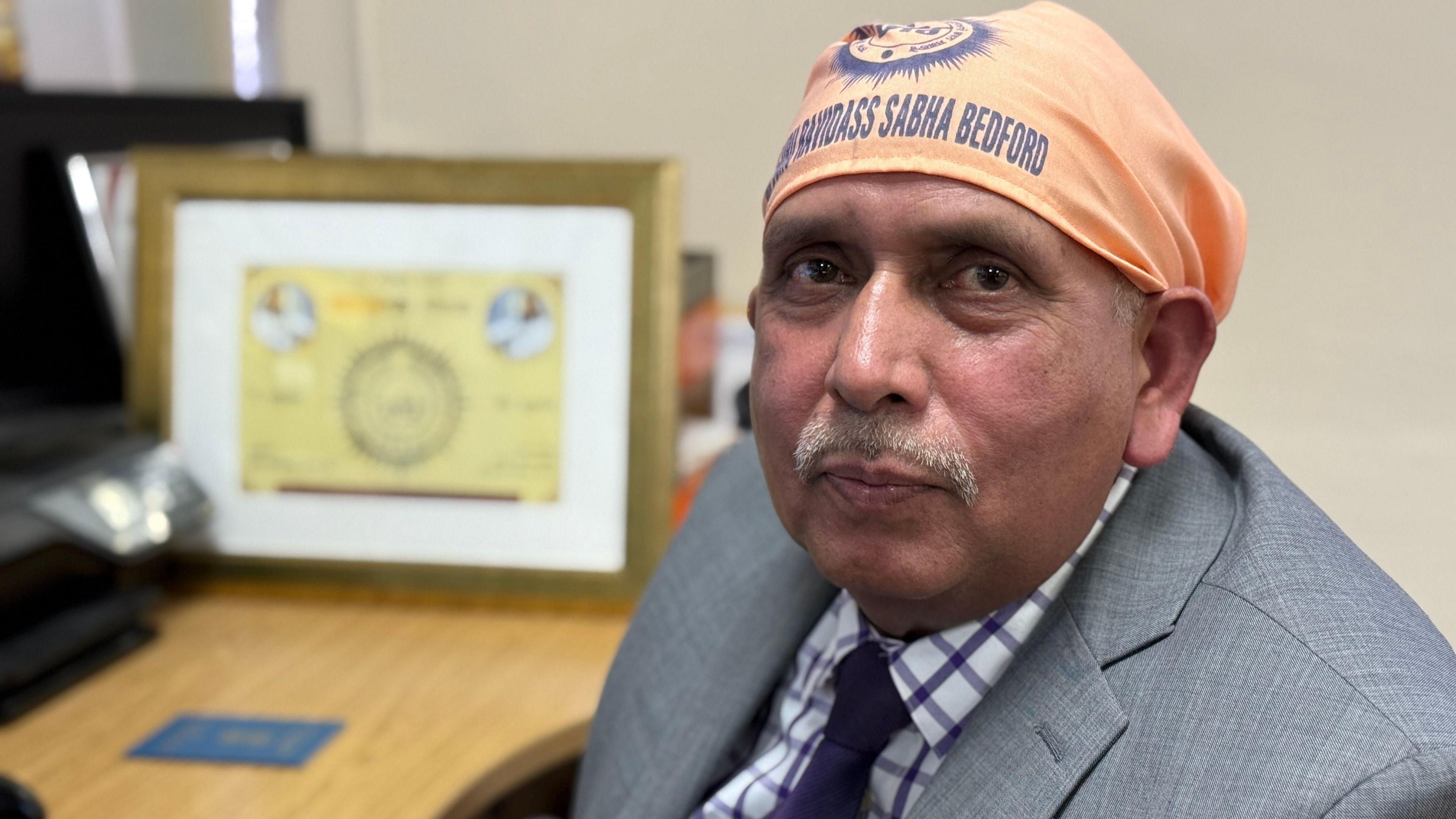 Jaswinder Kumar sitting down at a desk. He is wearing a grey suit jacket with a white and purple checked tie. He is looking at the camera. 