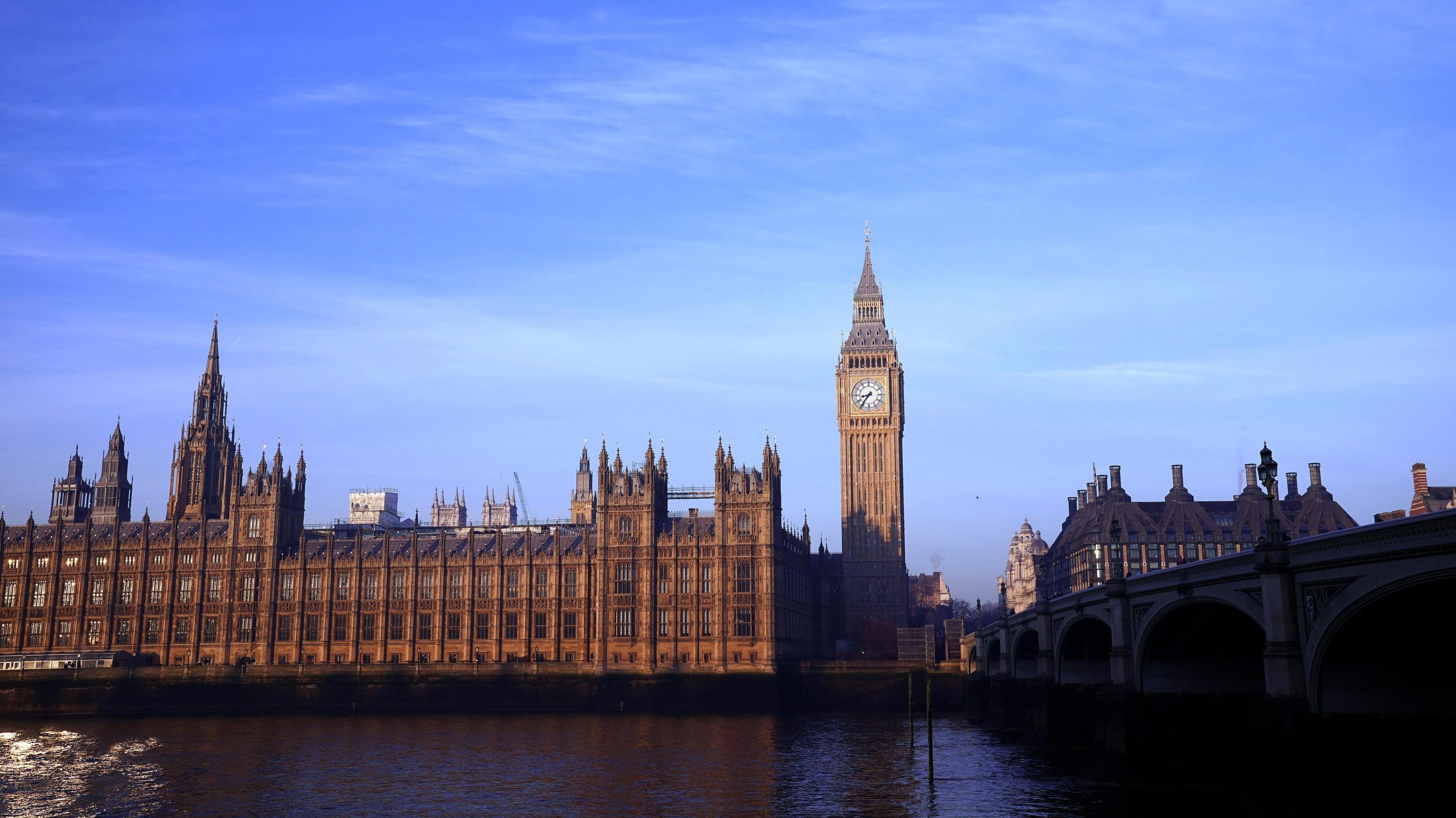 London's Houses of Parliament is lit up by the sunlight, against a backdrop of a blue sky. The River Thames and Westminster Bridge remain in the shadow. The time on Big Ben reads 8:35. 