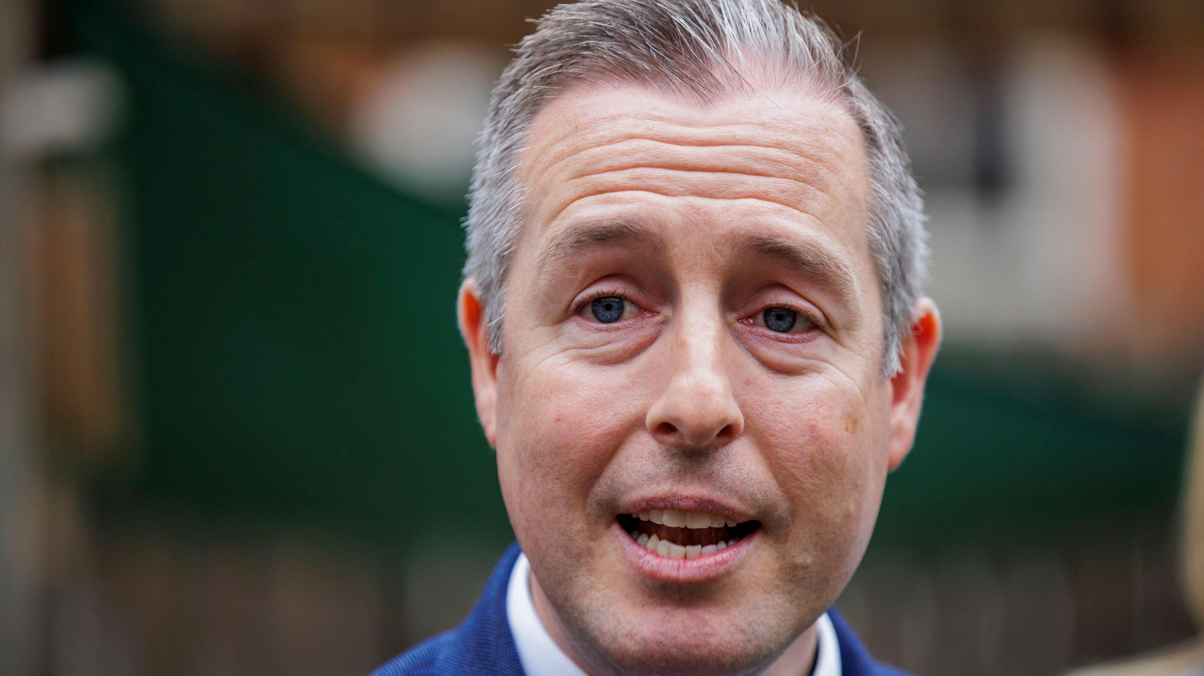 Paul Givan is pictured from the shoulders up. He has grey hair and is wearing a blue suit jacket and a white collared shirt. The background is blurred and he is mid-speech.