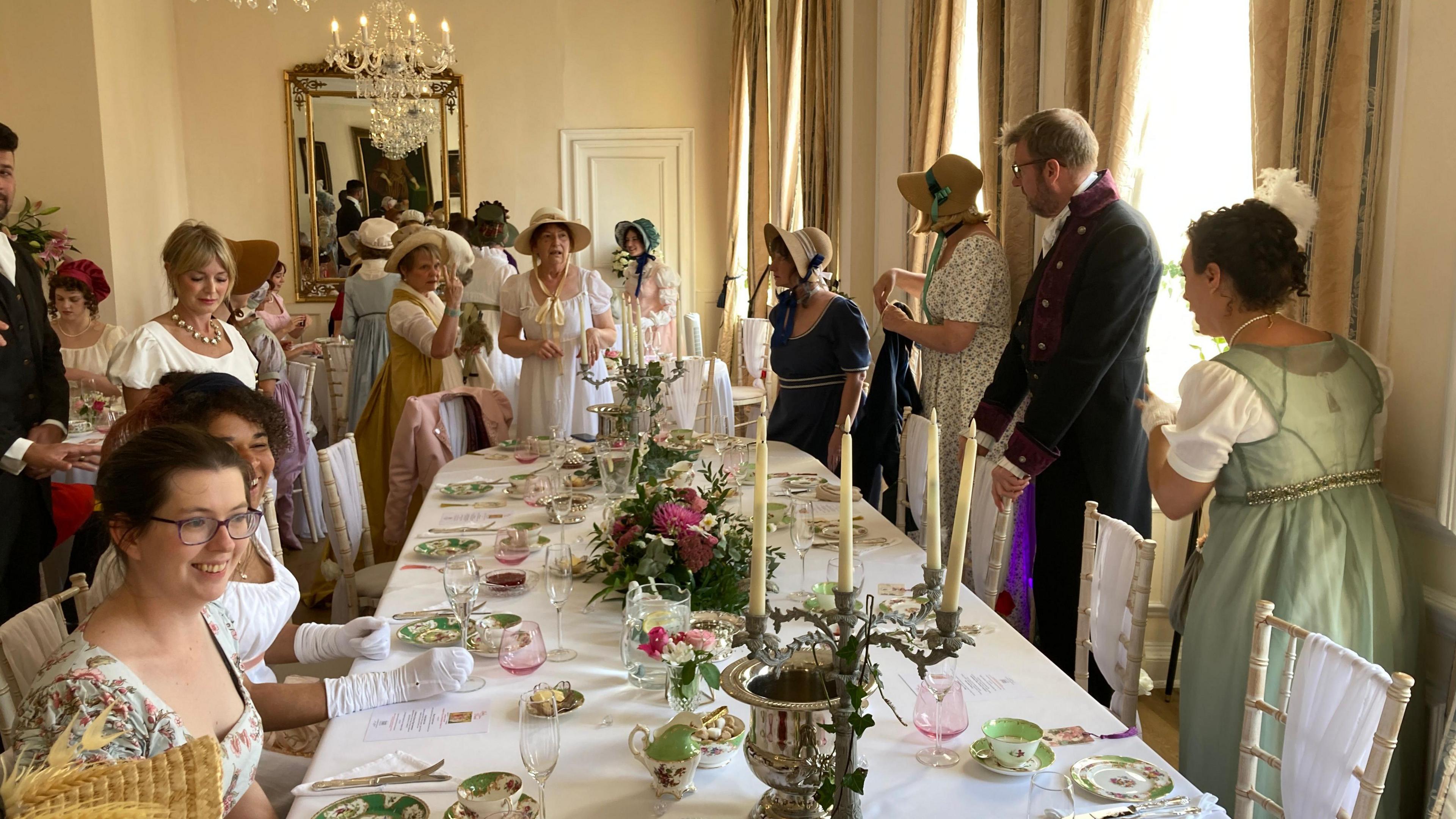 Around 20 people standing and sitting around a table in a ballroom in Parade House. The table is set with candlesticks, champagne glasses, and delicate china teacups. There is a large gold mirror on the wall and a glass chandelier hanging from the ceiling. Everyone is dressed in period clothing in light pastel colours. 