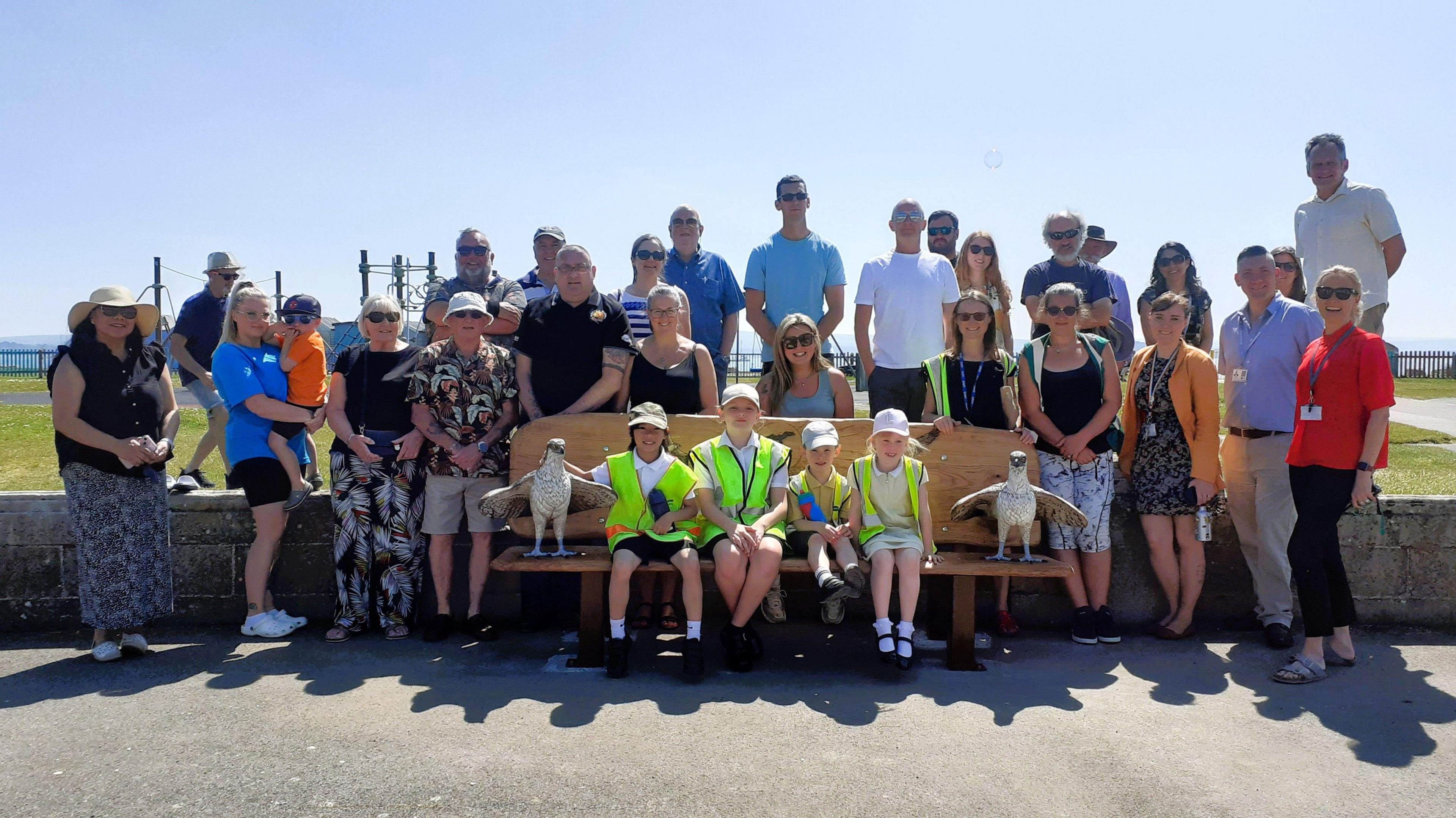A large group of people, including four primary school children wearing high-visibility jackets, smiling around a wooden bench with a carving of an osprey on each end. It's a sunny day and the sky is blue.
