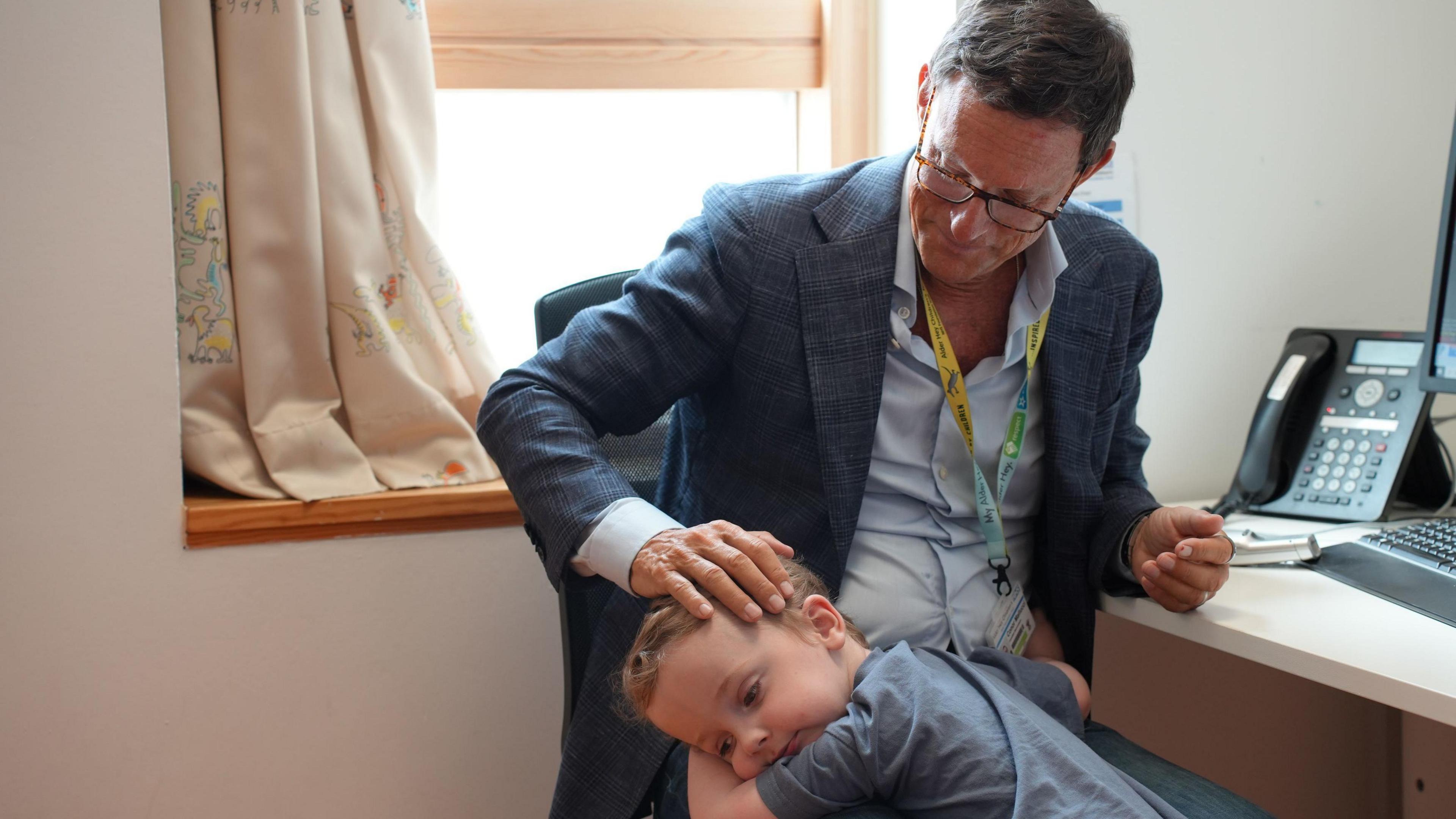 Conor, now three, from Bolton, hugging his surgeon Conor Mallucci in his office. The surgeon apts his head as he sits at his desk with a phone and keyboard on it.