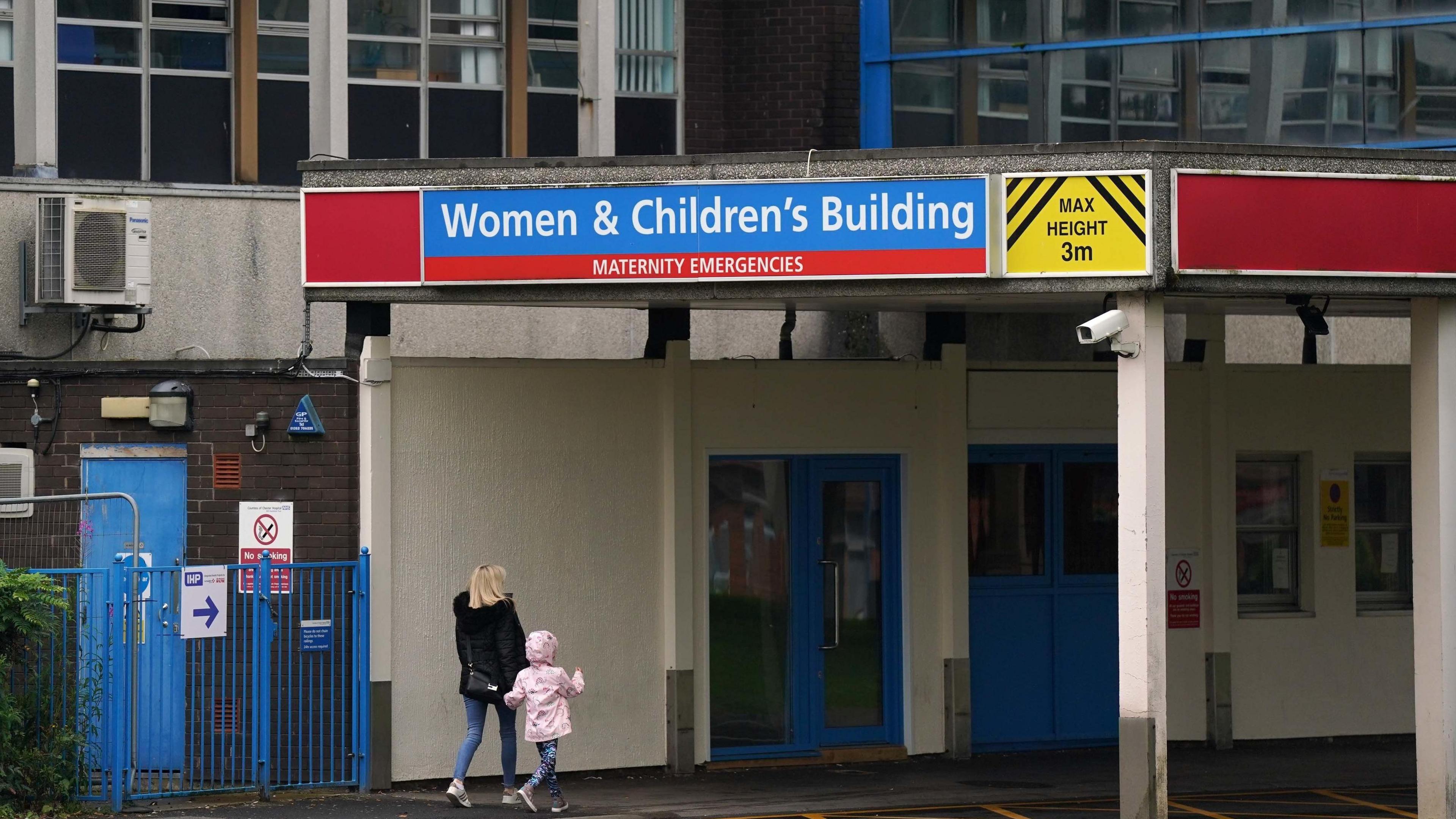 An unidentified blonde-haired woman wearing a black coat walks hand-in-hand with a young girl in a pink coat. They both have their backs to the camera as they make their way towards the entrance of the Women & Children's Building at the Countess of Chester Hospital, which is marked with a large blue sign.