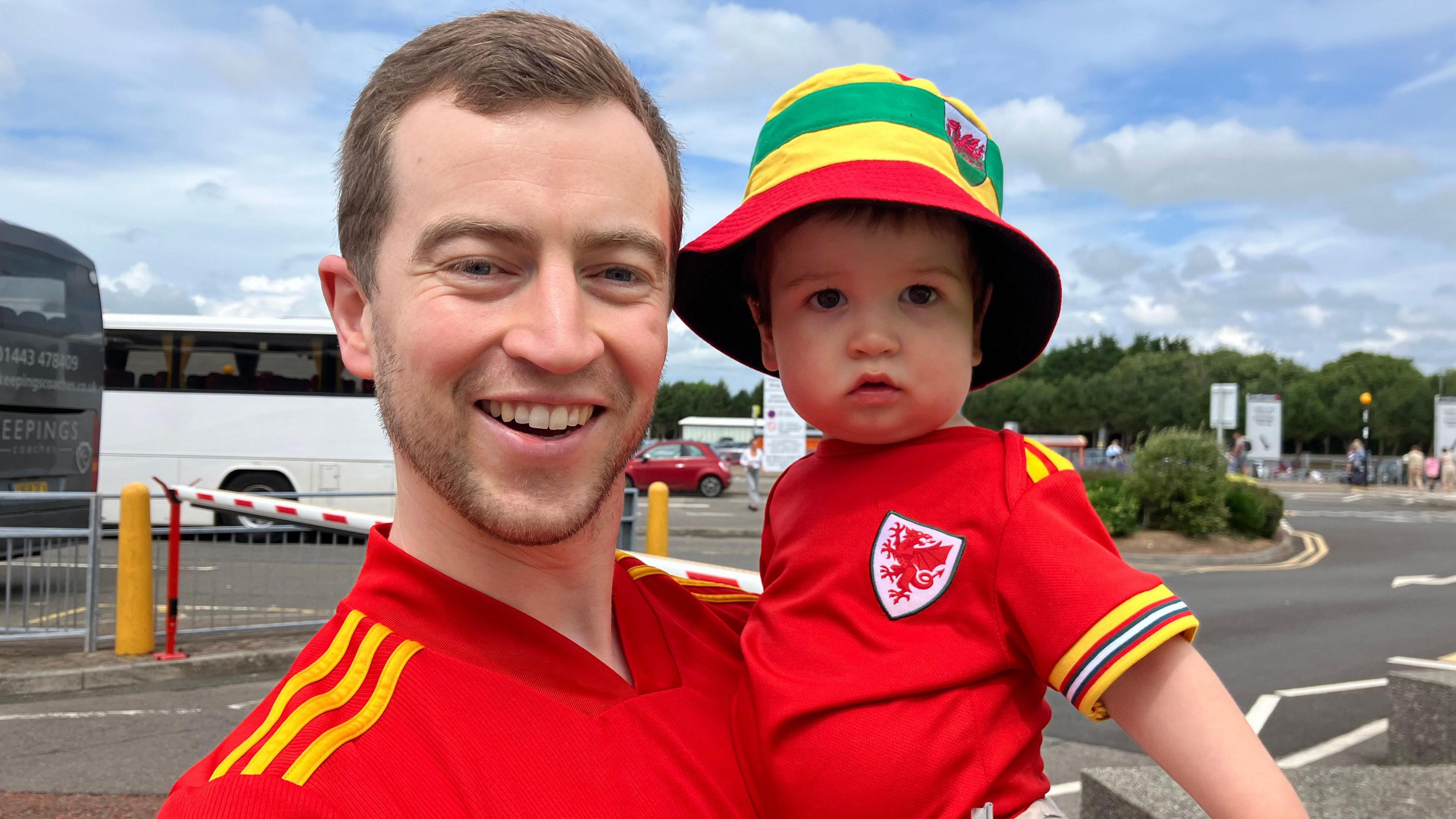 A man with short brown hair holds his baby son, who has a red, yellow and green hat. Both are wearing red Wales football shirts.