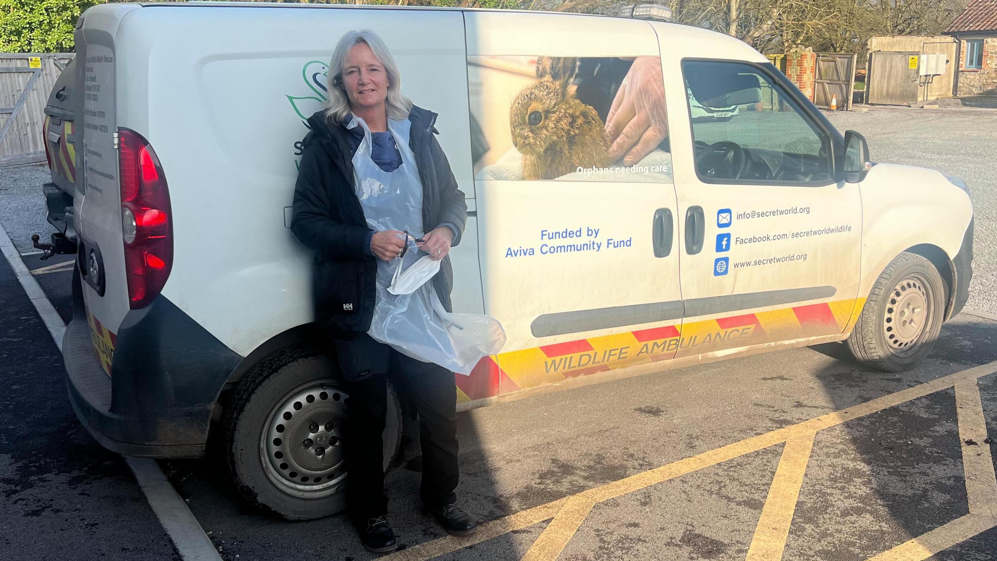 Volunteer driver Tracy Abbot is standing next to a white van used to transport animals. She is wearing a black coat and plastic protective wear. The van says 'Wildlife Ambulance' on the bottom.