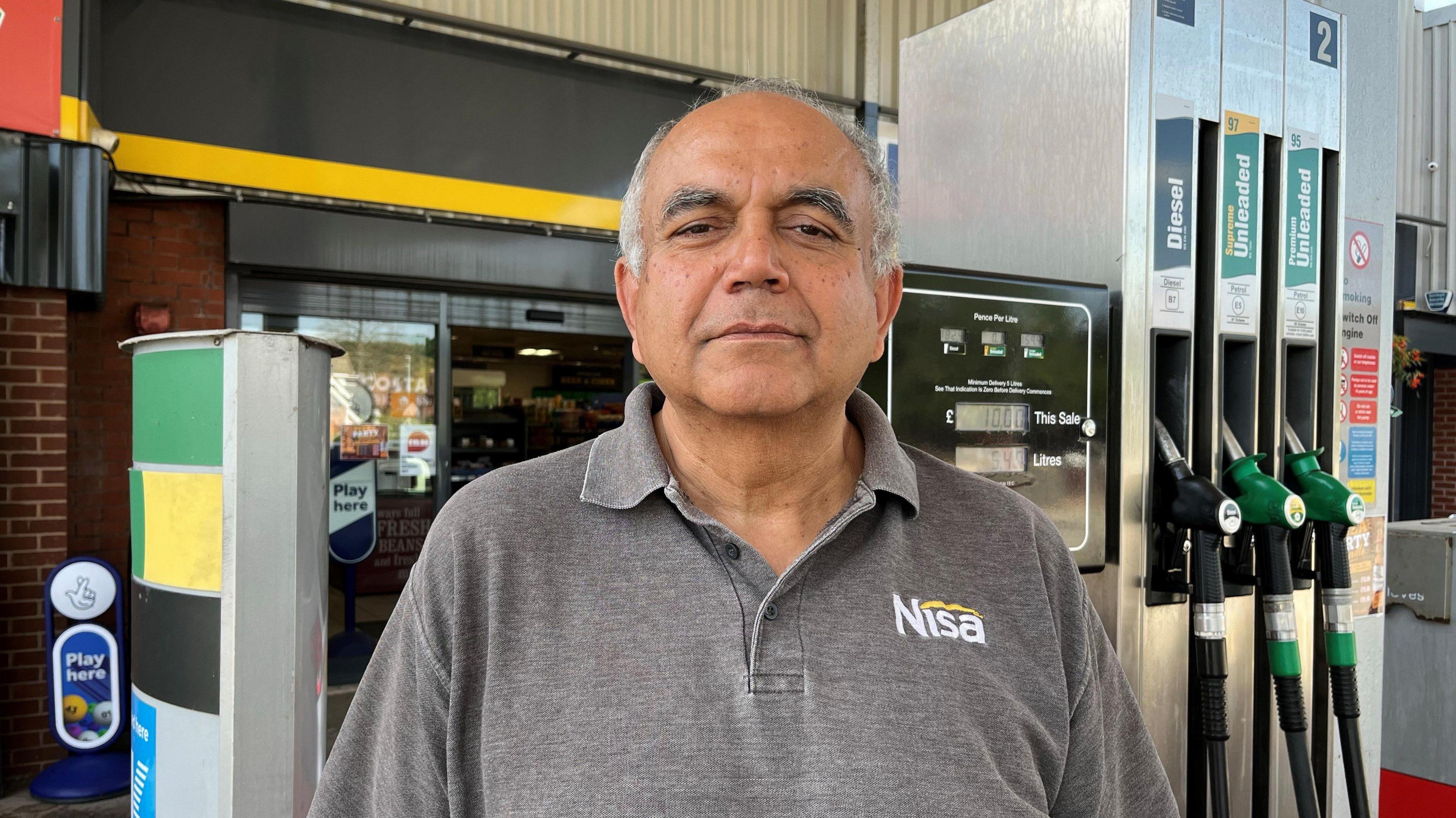 Shailesh Parekh is wearing a grey polo t-shirt with the Nisa logo on it as he stands in front of the fuel pumps on one of his forecourts.