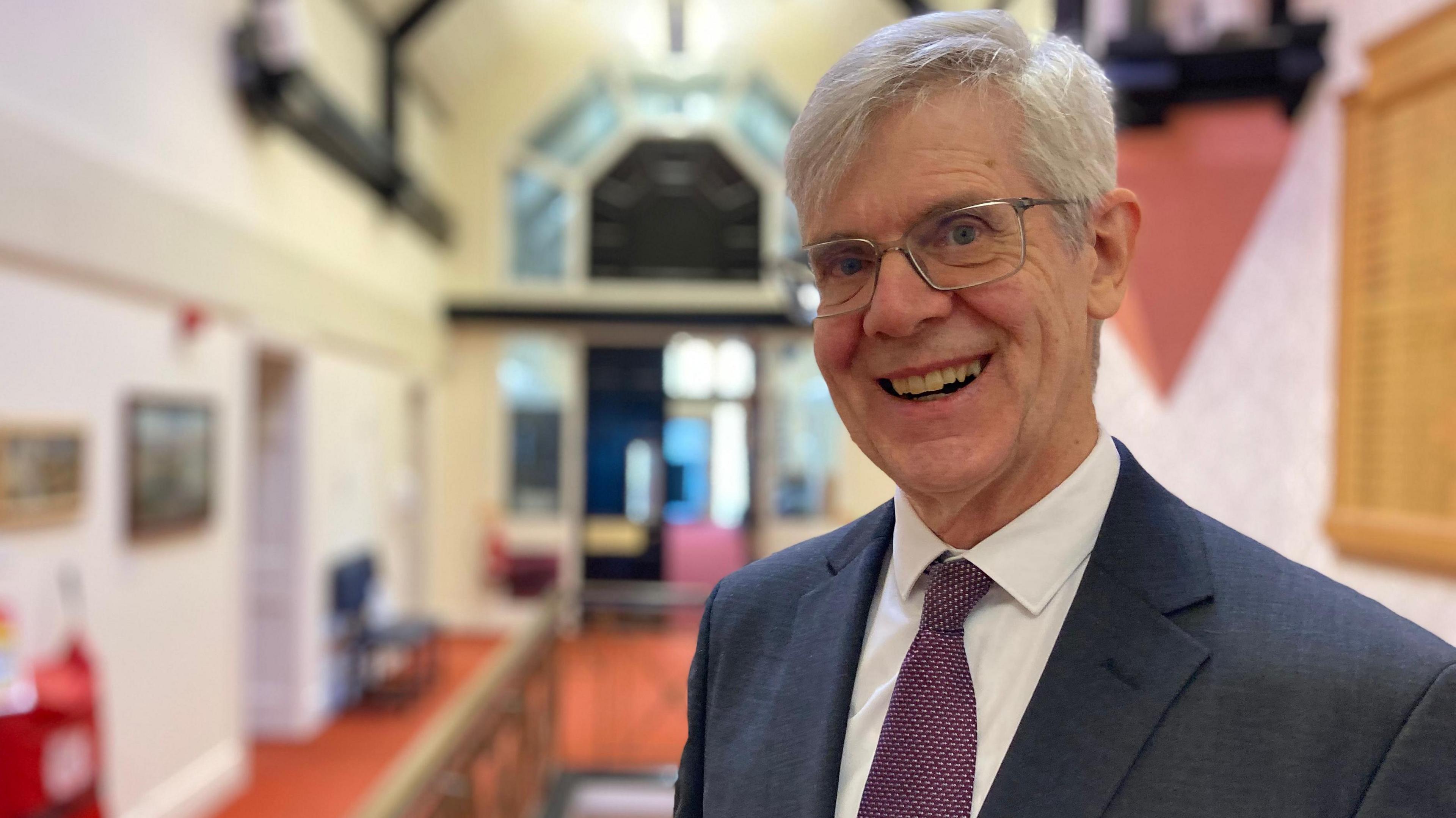 Llun o ddyn yn sefyll mewn coridor swyddfa'r cyngor yn gwenu at y camera. Mae ganddo wallt llwyd a sbectol. Mae'n gwisgo siwt llwyd tywyll, crys gwyn a thei porffor patrymog. 