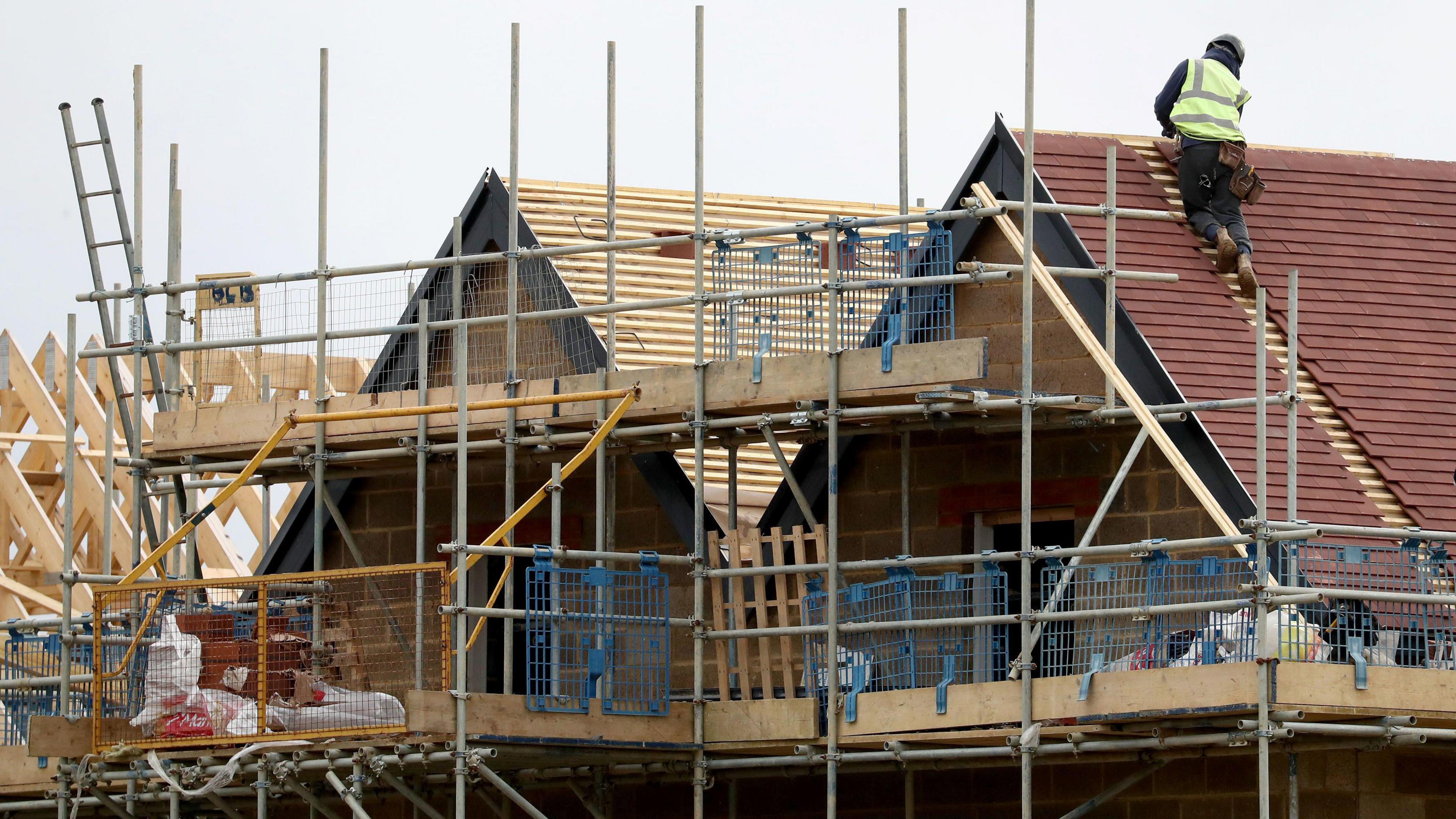 A builder on the roof of a partially built house with scaffolding in the front.