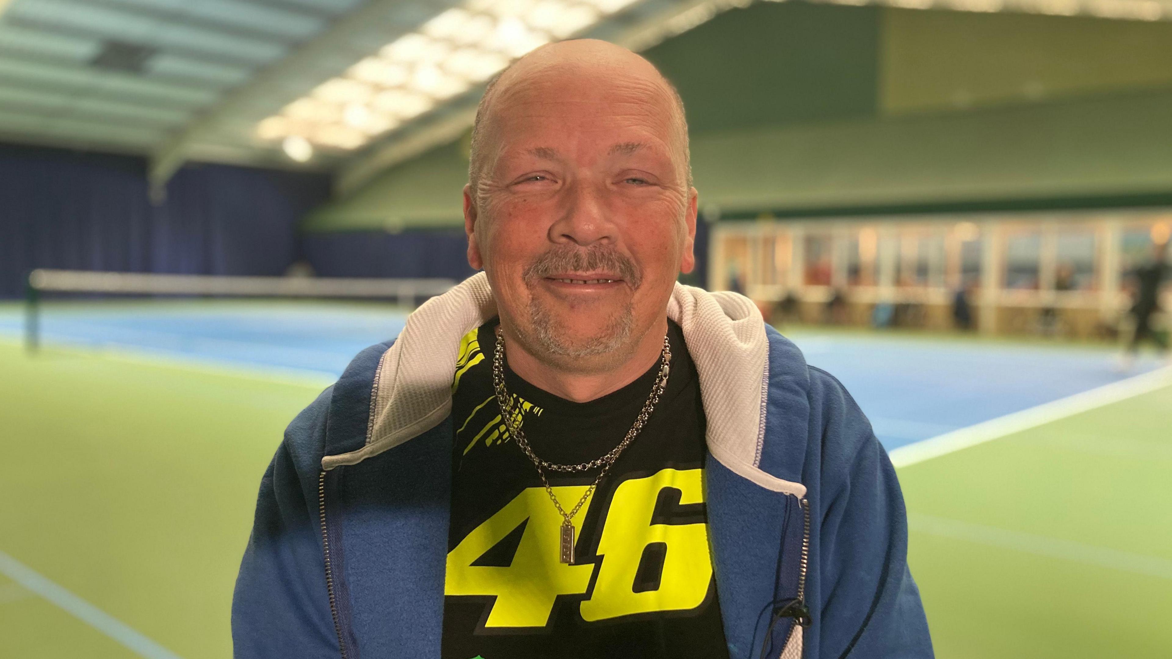 Neil wears a T-shirt with a big yellow 46 on it and a blue hoodie. He is on the side of a tennis court and smiles for the camera.