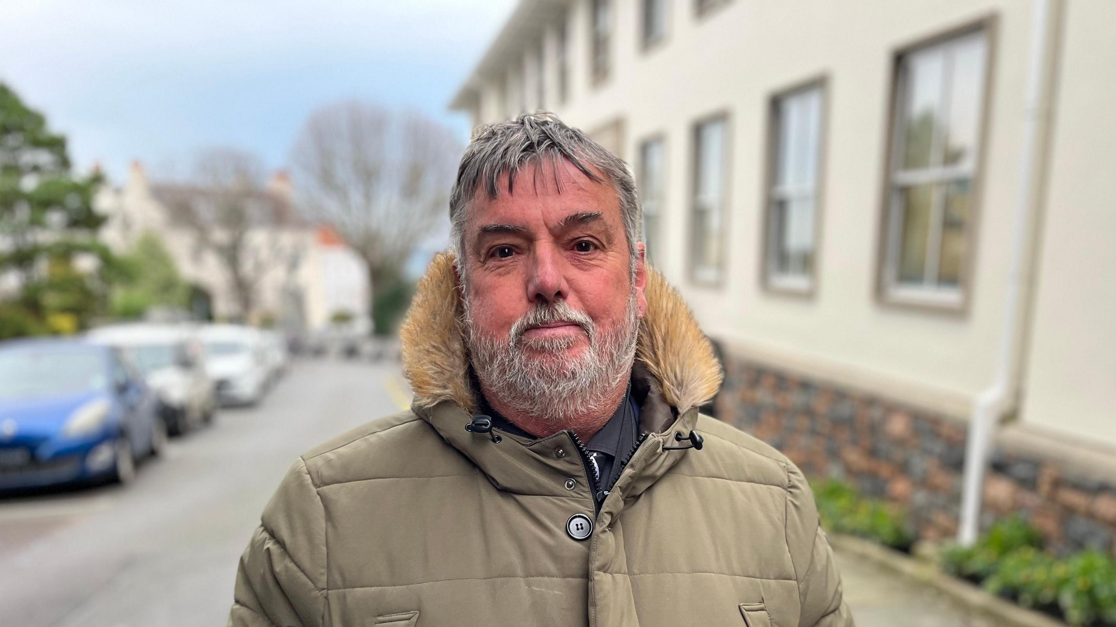 Deputy Peter Roffey wearing a khaki coloured puffer coat with a brown fluffy hood. He as dark grey hair and a grey beard. He is stood on a street with a cream coloured building over his left shoulder and a row of cars to his right.