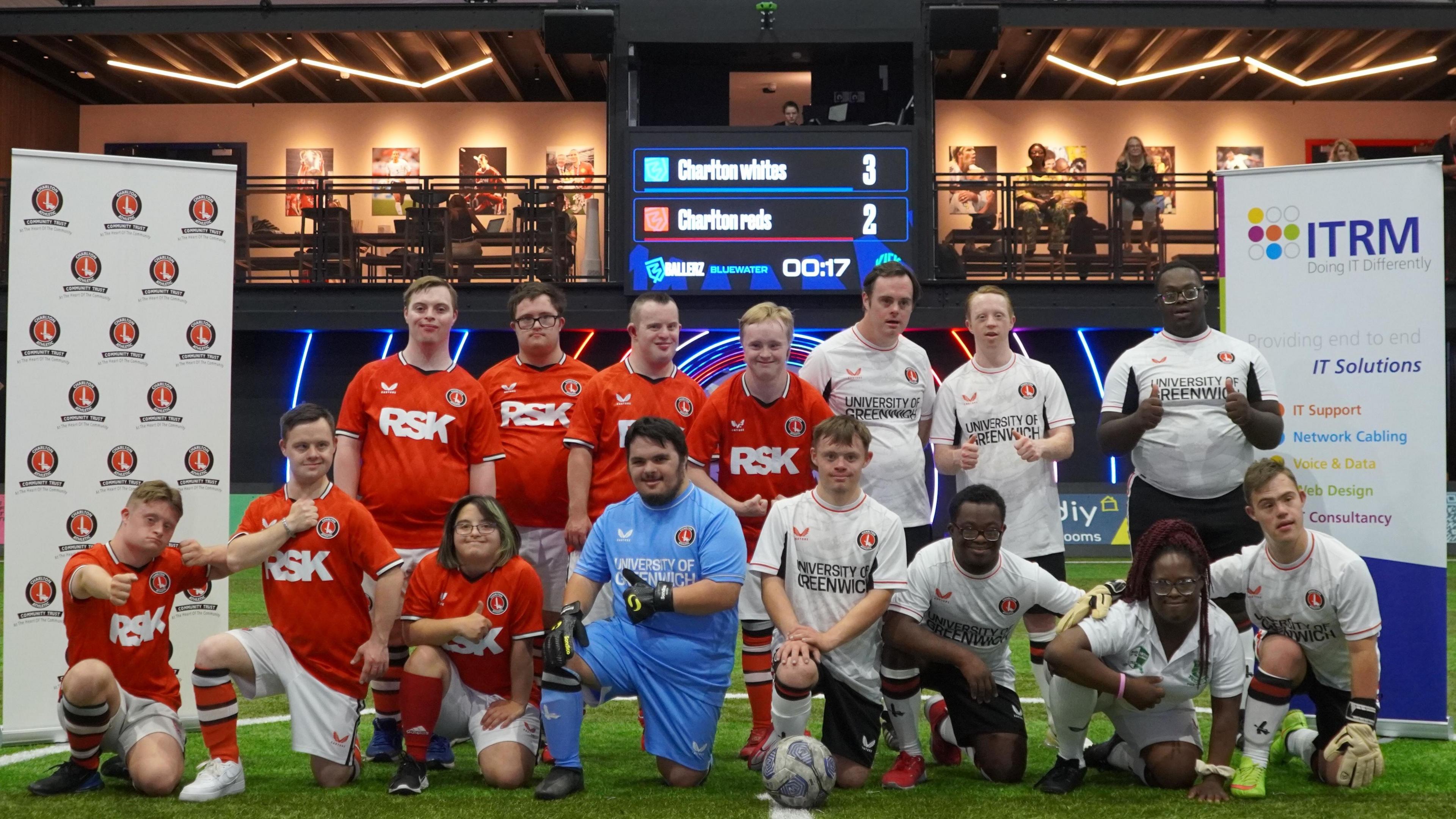 Fifteen football players posing on a pitch, with eight of them kneeling, and a football is central and in front of one of the players. Seven are wearing red football shirts with white shorts, and seven are wearing white shirts with black shorts. The one other play is wearing a blue shirt and blue shorts