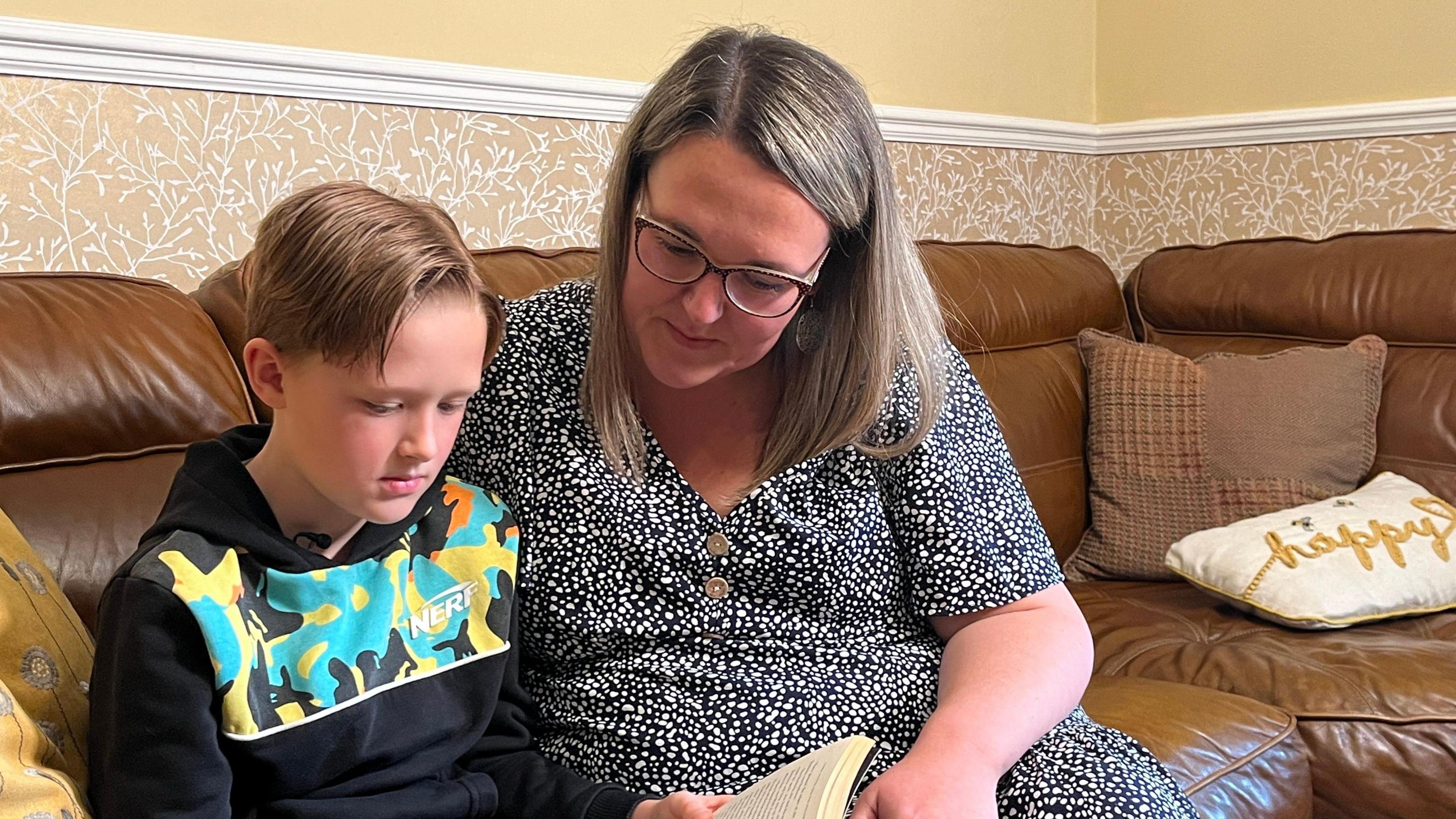 David reads a book sat beside his mum who has her arm around his shoulder.   