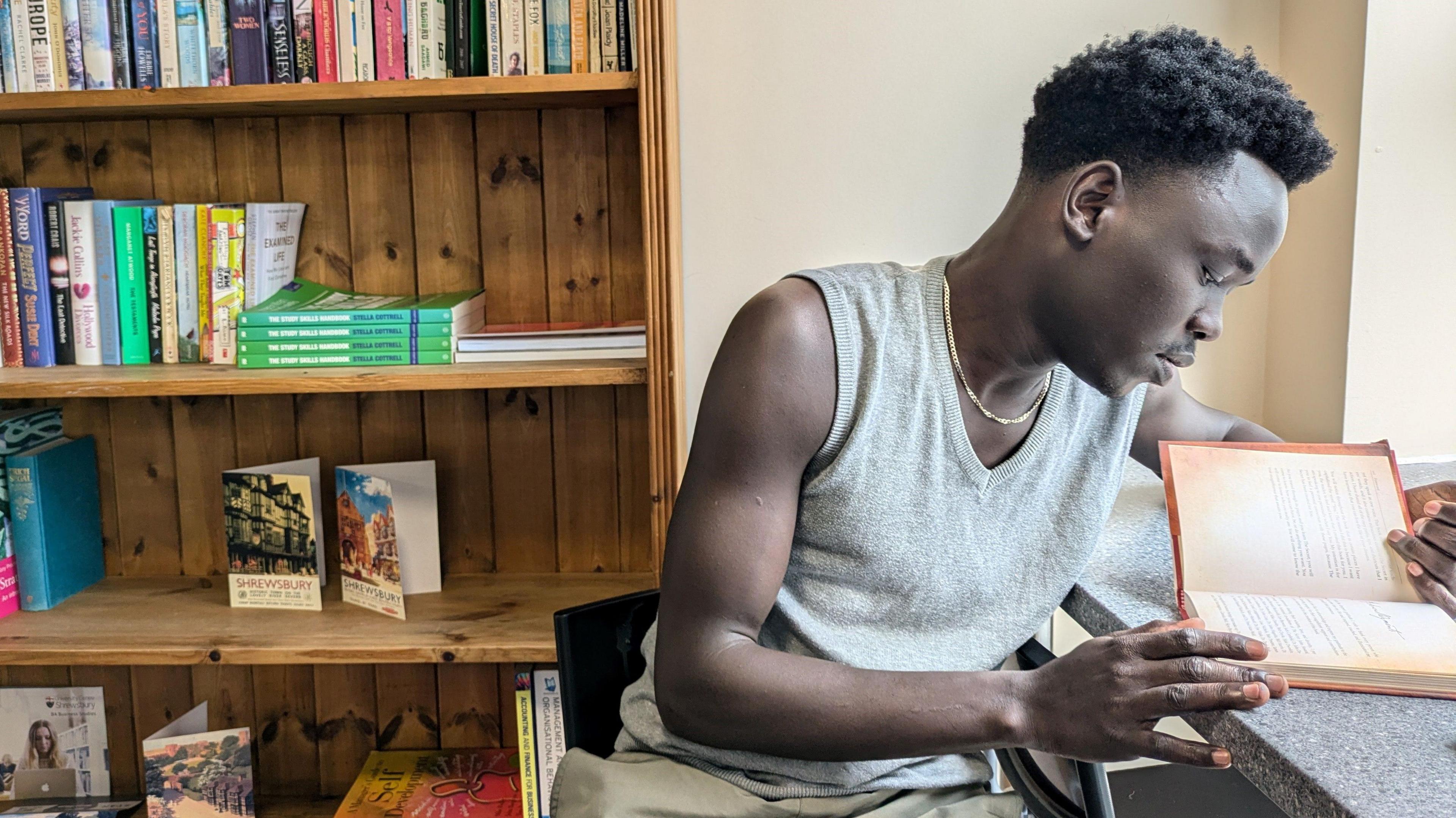 A man sits facing to the right of the frame and looks down at an open book on a desk. He wears a grey, sleeveless top and has a thin gold chain around his neck. He sits on a chair facing away from the desk and behind him is a wooden bookshelf with several books on it.