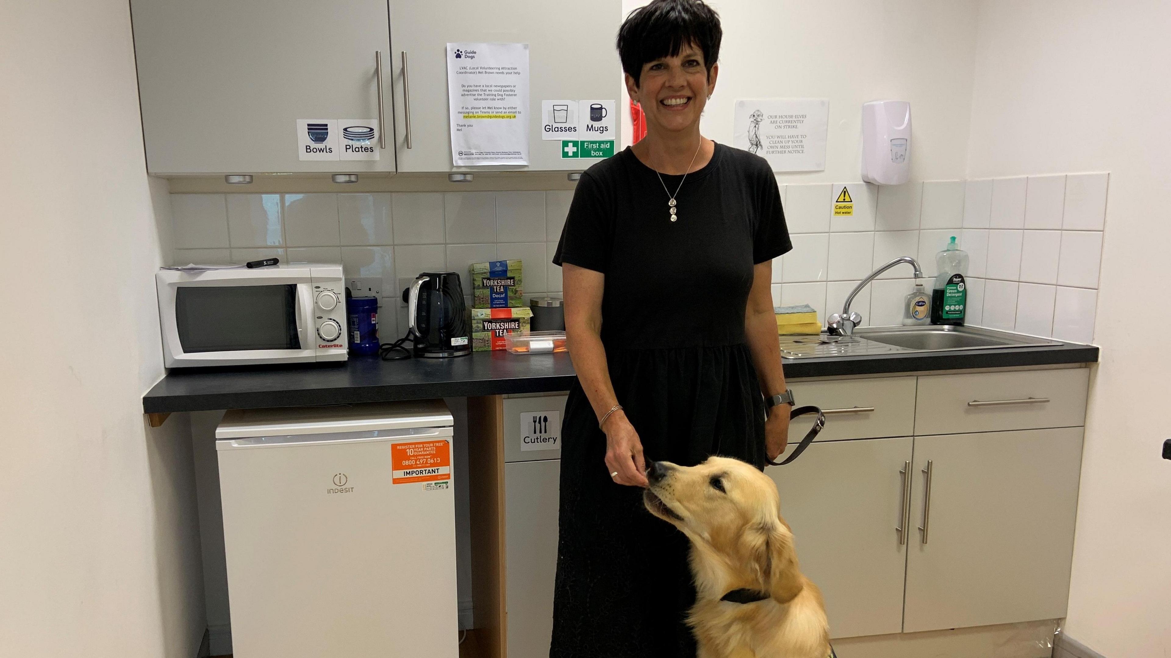 A woman with short black hair is wearing a long black dress. She is standing in a kitchen with a golden labrador guide dog sitting upright beside her