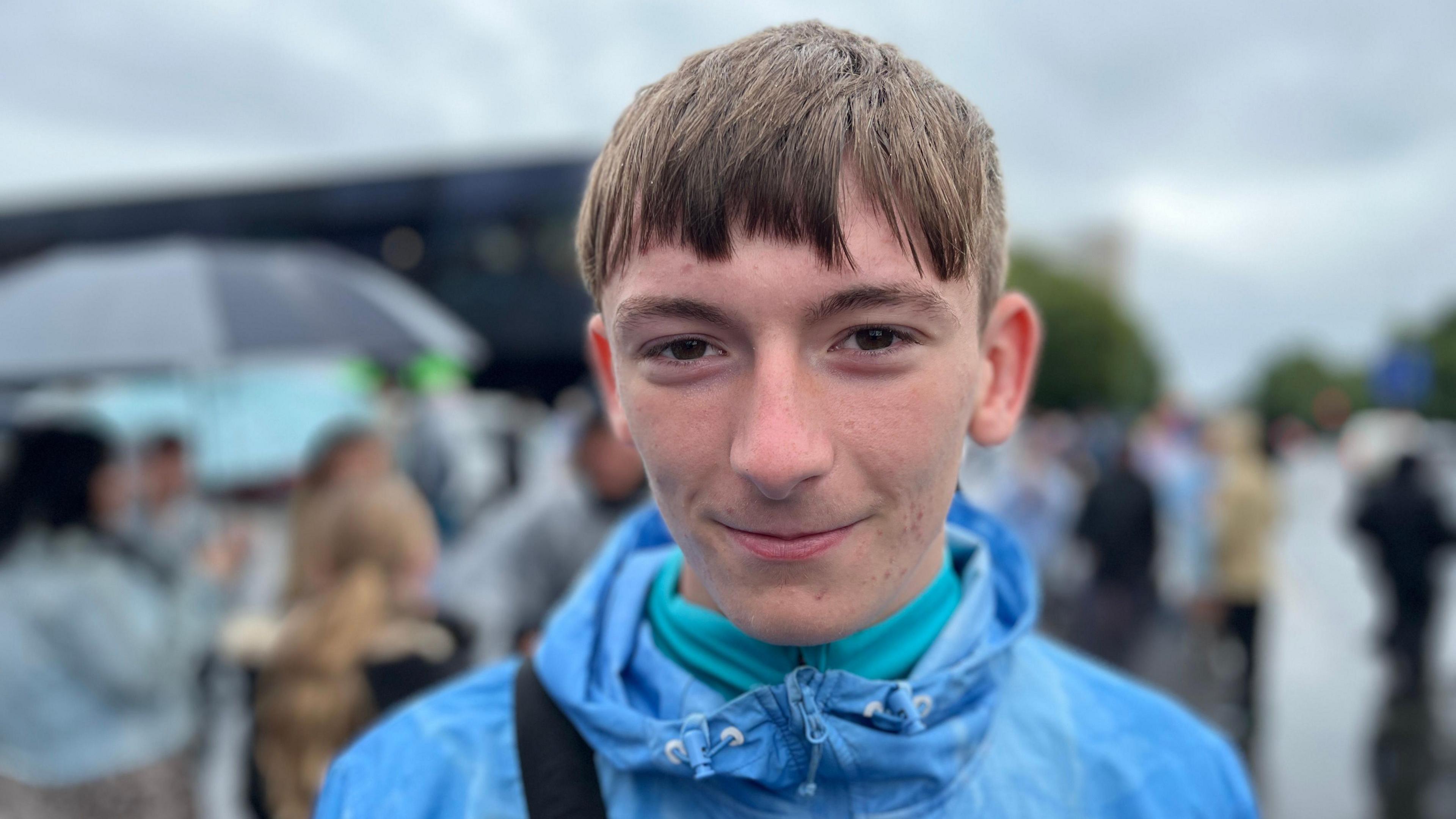 A boy looking at the camera. He has dark hair and dark eyes. He is wearing a light blue jacket with a turquoise jumper underneath. The background is blurred and the sky is grey.
