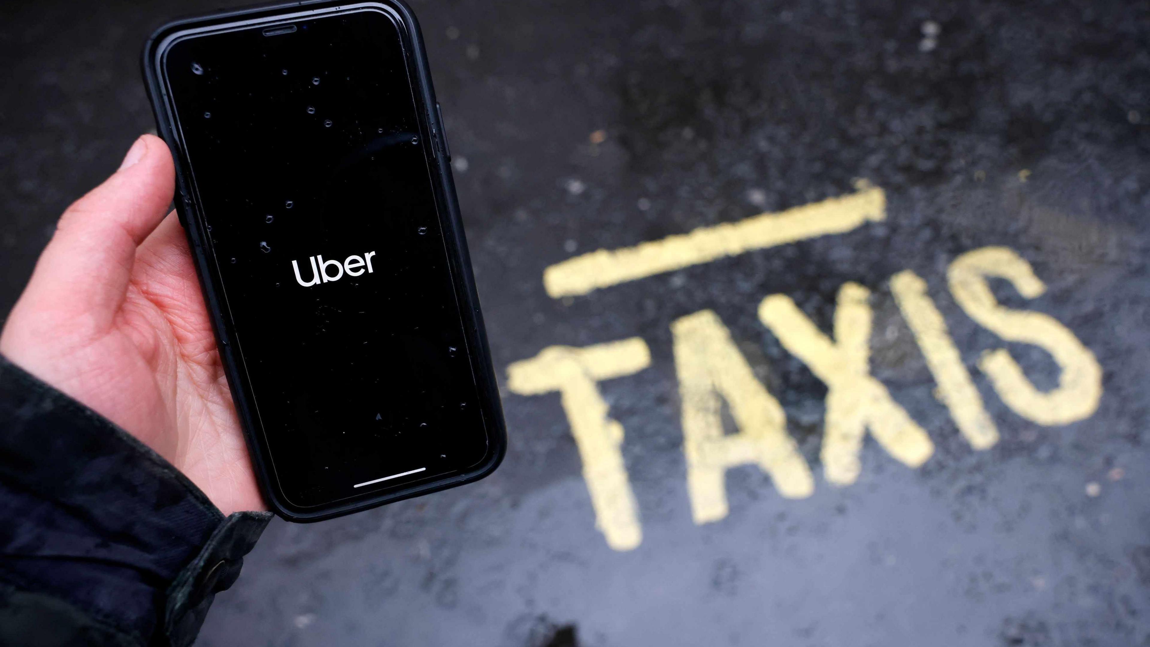 A person holding an apple i-phone with the uber app open, standing next to a yellow sign saying "taxi" on the road.