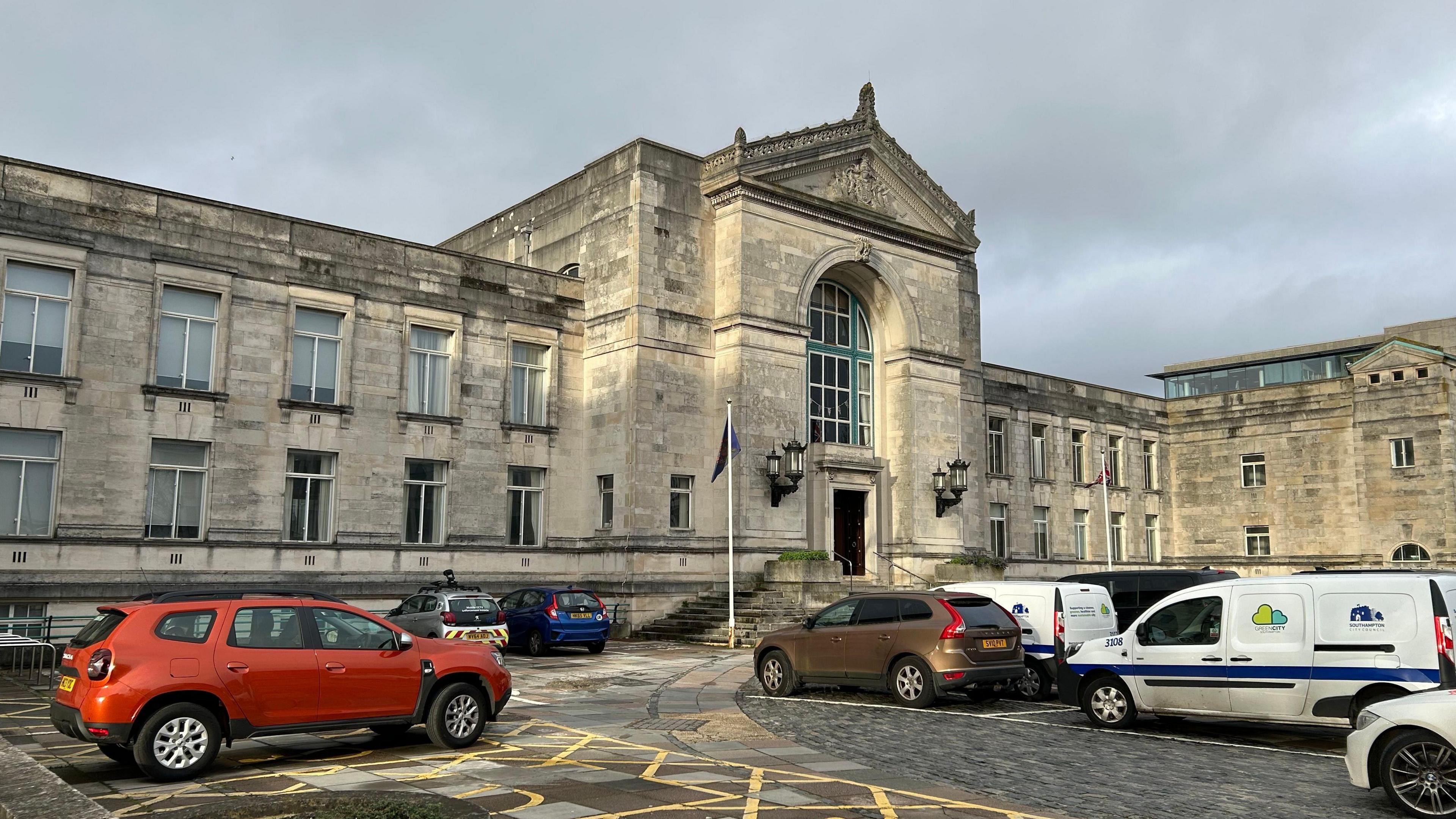 Exterior view of Southampton Civic Centre, a large cream brick building with blue detailing on am arched window at the entrance. Can also see lots of cars parked outside and a flagpole at the base of the steps to enter the building.