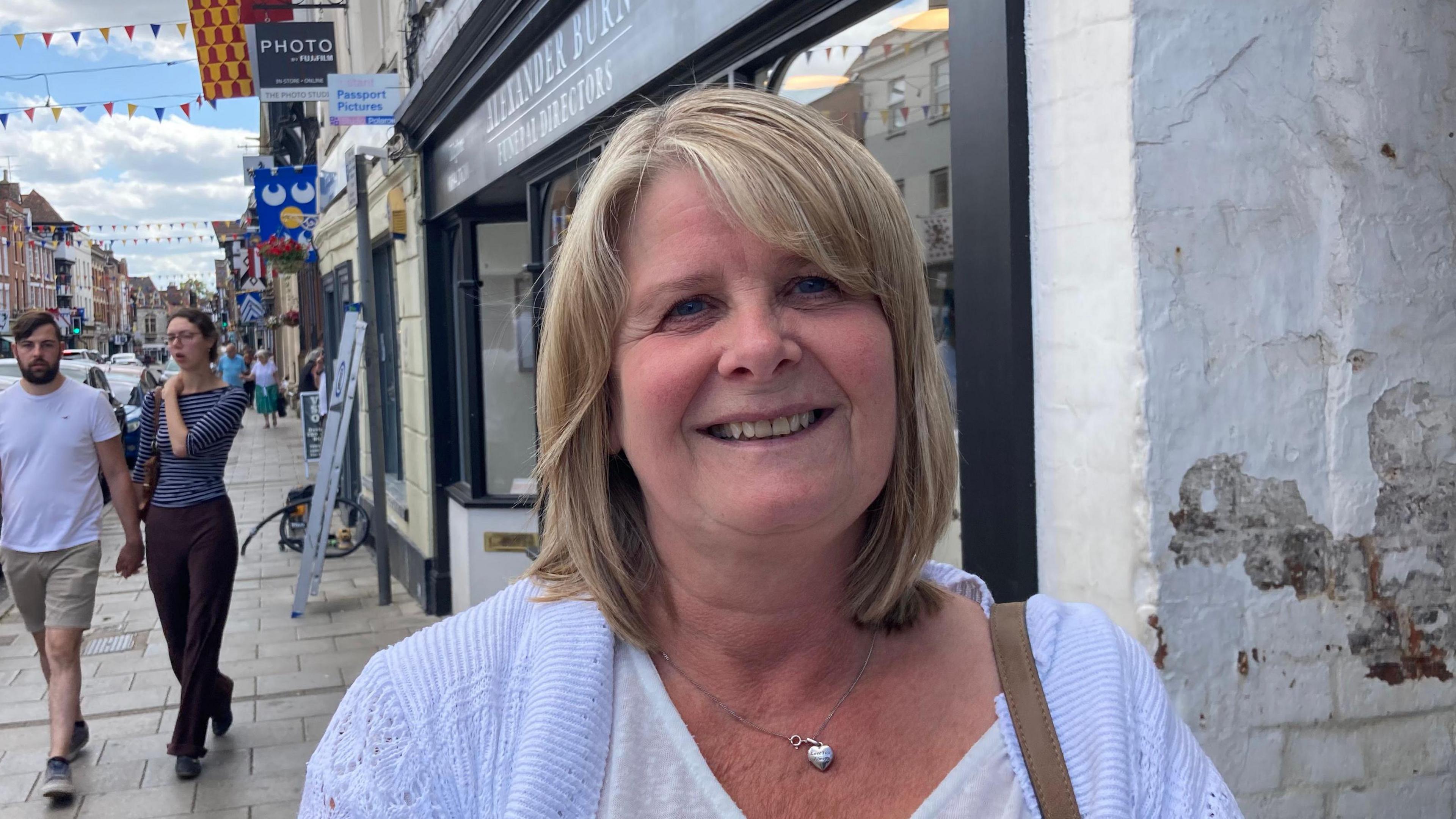 A photo showing a woman with blonde hair. She is wearing a white top and light blue cardigan, she is also wearing a heart shaped necklace. She is stood in front of a shop front with a man and woman walking past in the background