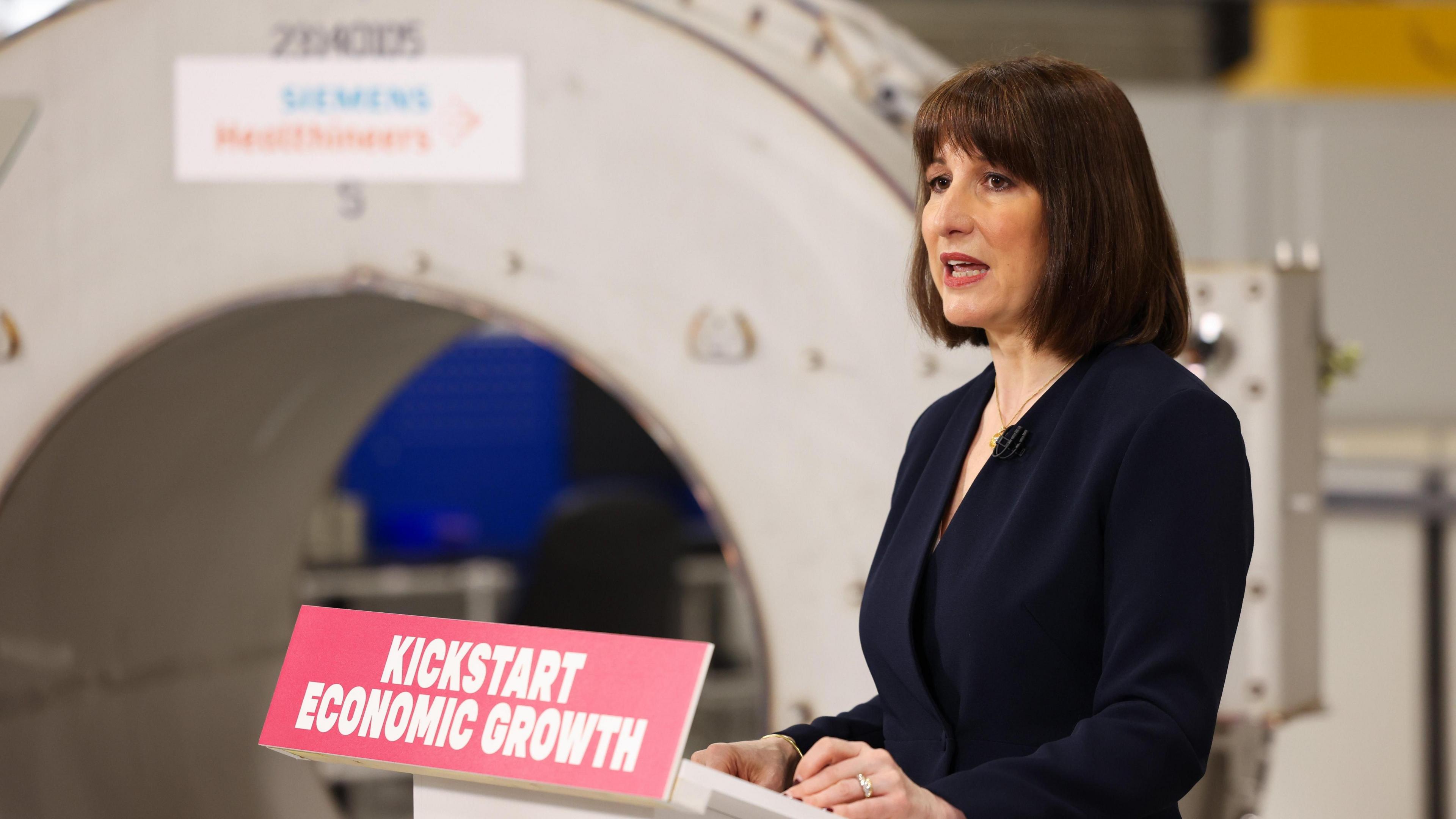 Rachel Reeves delivers a speech on UK growth at the Siemens Healthineers factory near Oxford. On her plinth is a sign that says 'Kickstart Economic Growth'. She has short dark brown hair and wears a dark blue blazer. In the background is a large circular piece of scientific equipment.