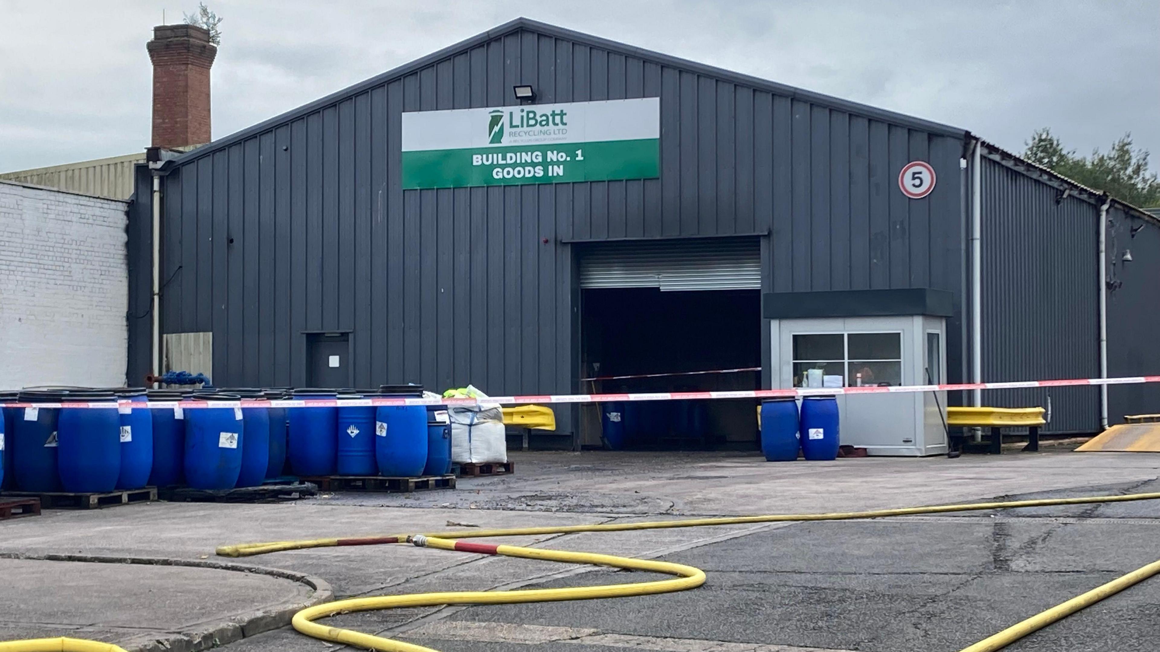 A factory cordoned off with tape. The building says LiBatt and BUILDING No.1 GOODS IN in green letters